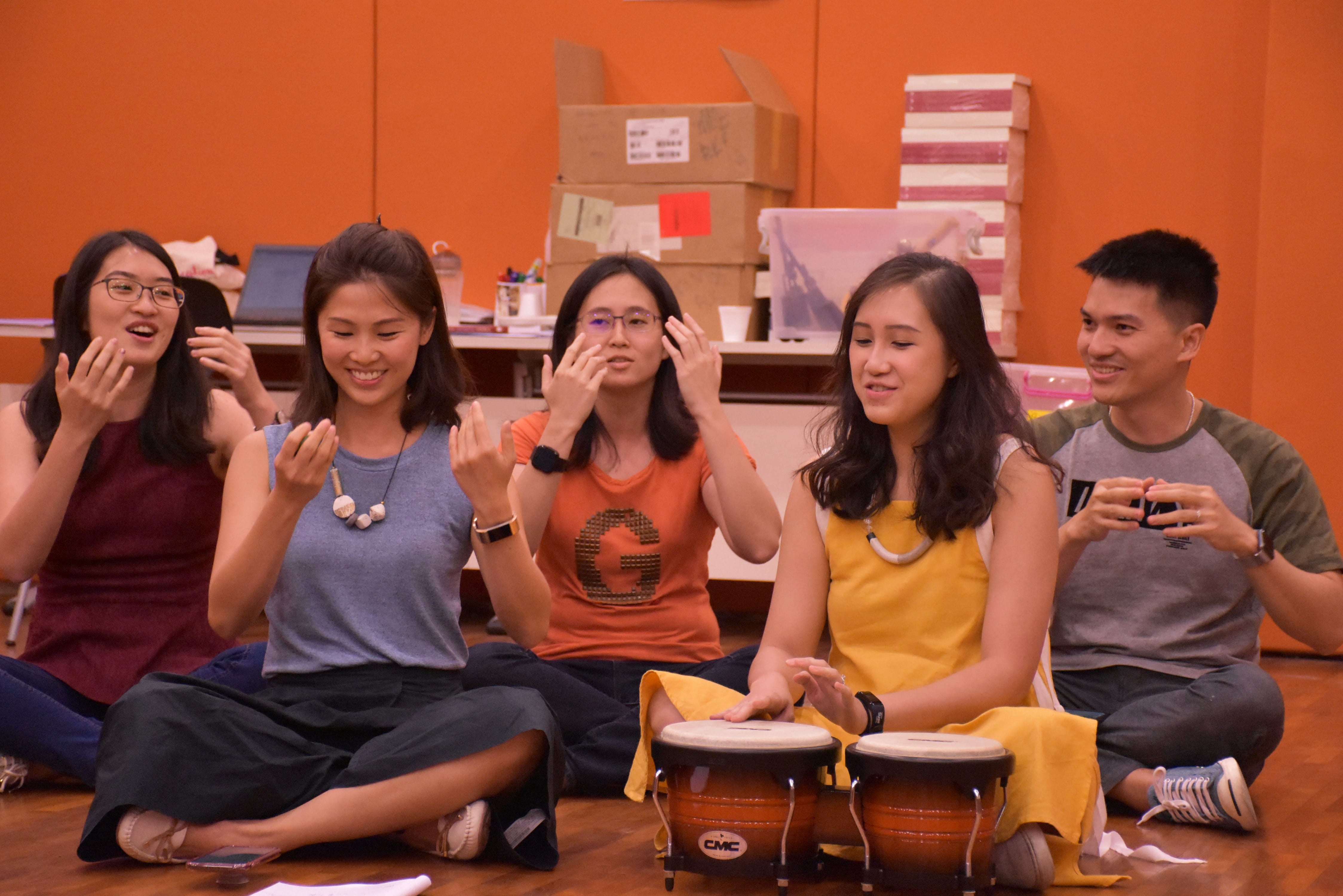 Photo taken during Teaching Living Legends worshop. Five teachers sit on a wooden floor smiling, one of them with bongo drums in front, the others doing actions and hand gestures, against an orange wall.