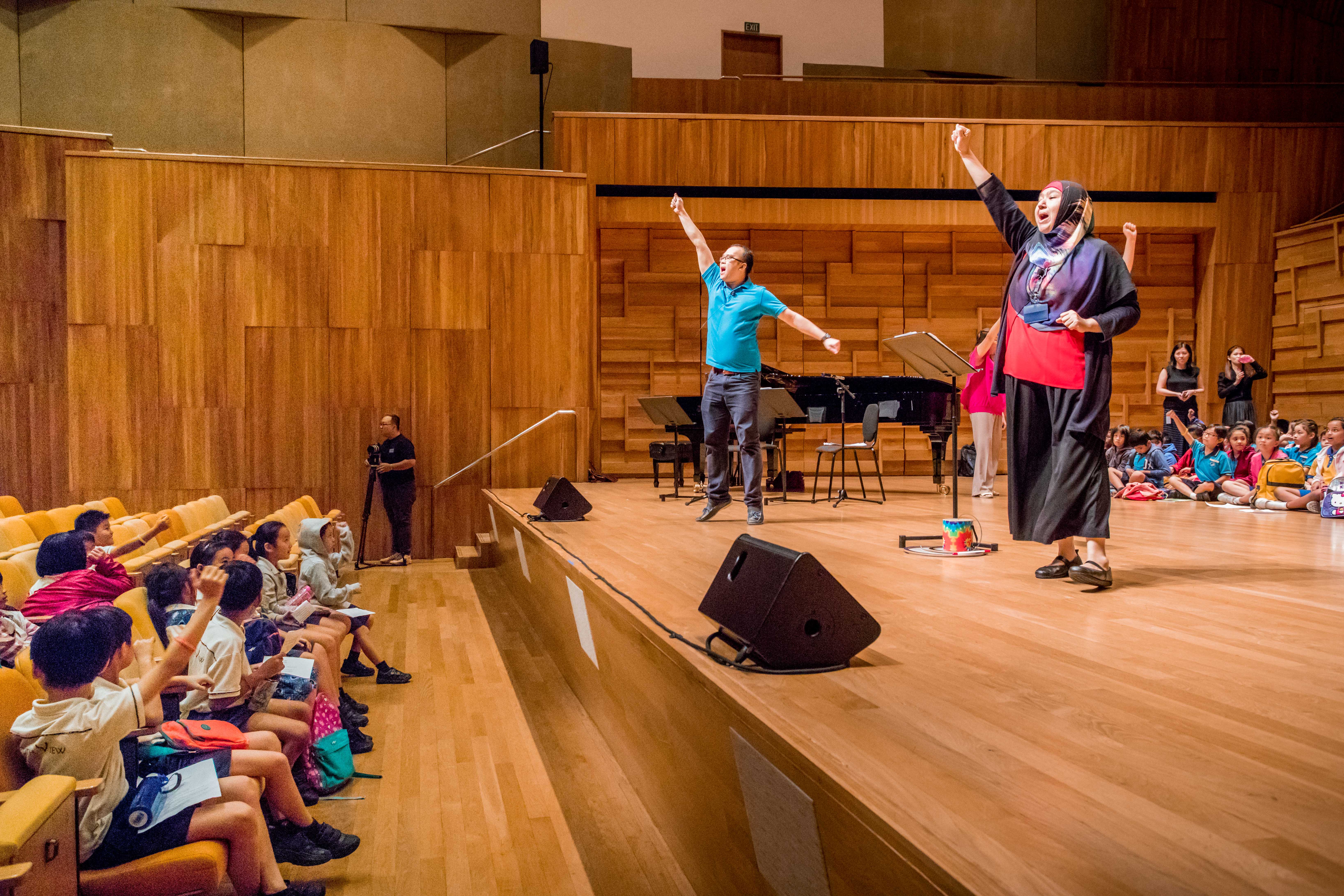 Photo taken during Engaging With Songs Day. Two facilitators raise their arms on a wooden concert hall stage while children follow the action from seats and the floor.