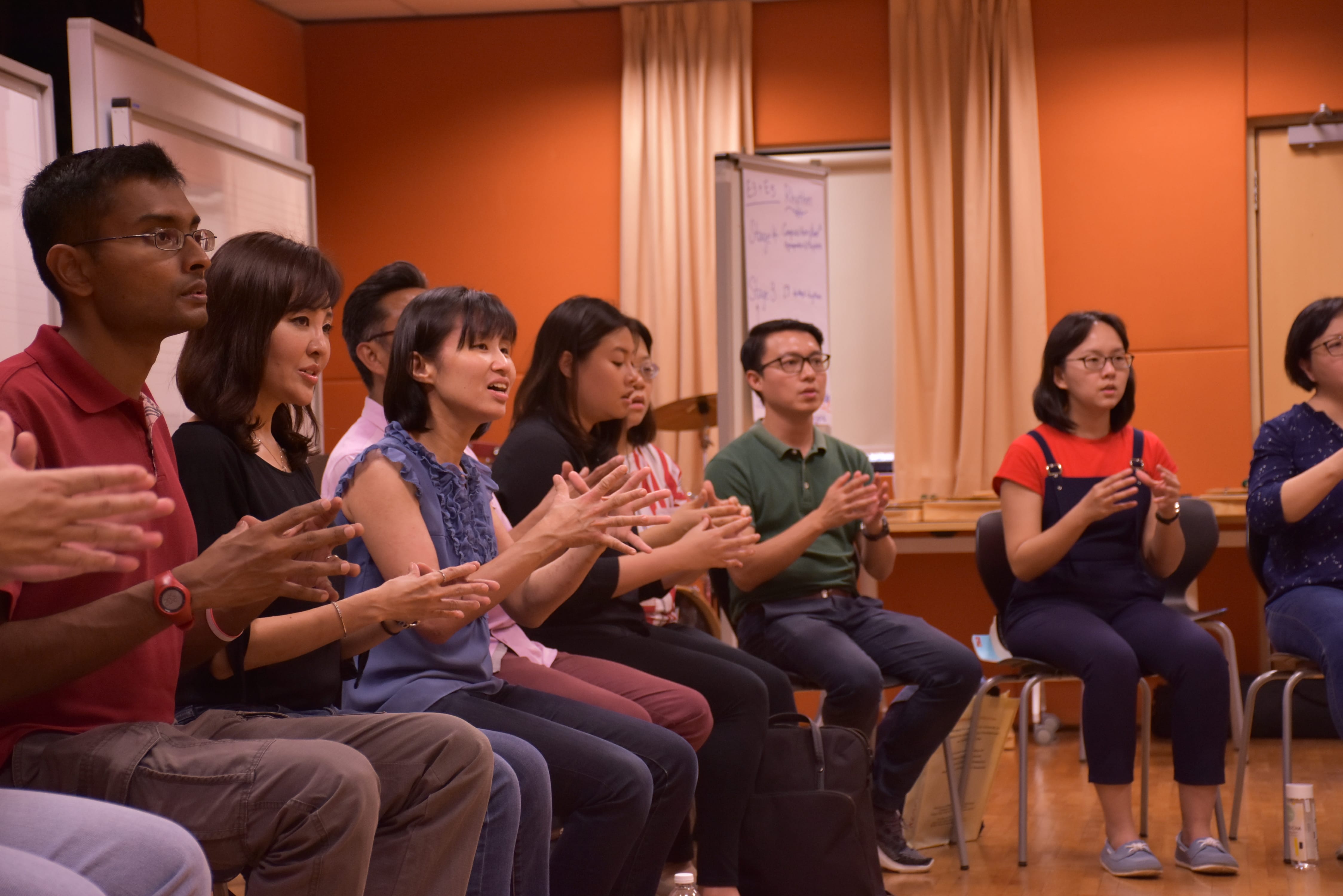 A group of music educators seated in a room with orange walls, clapping hands during a music activity in workshop.
