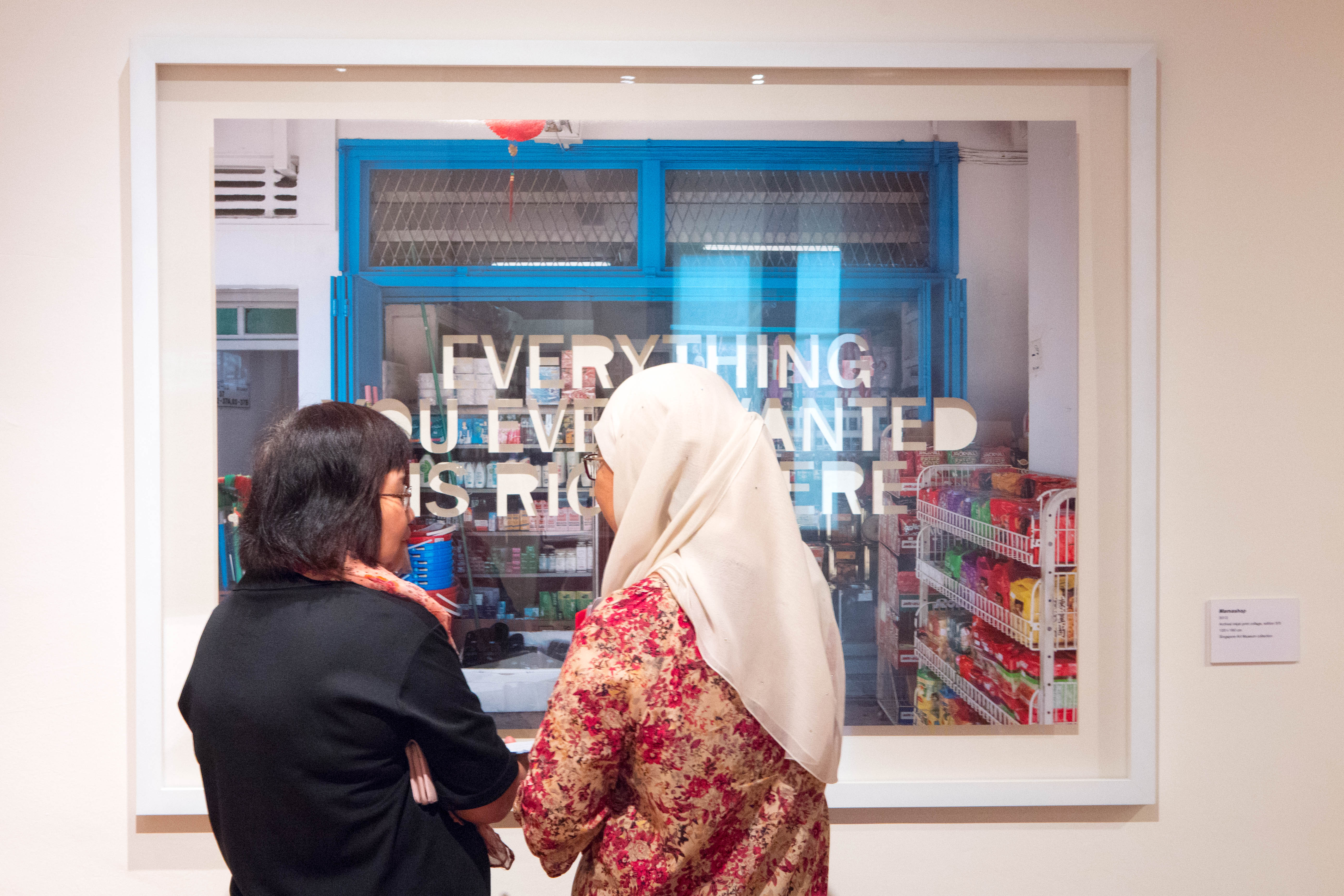 Two women view a framed artwork showing a shop with text reading "Everything You Ever Wanted Is Right Here."