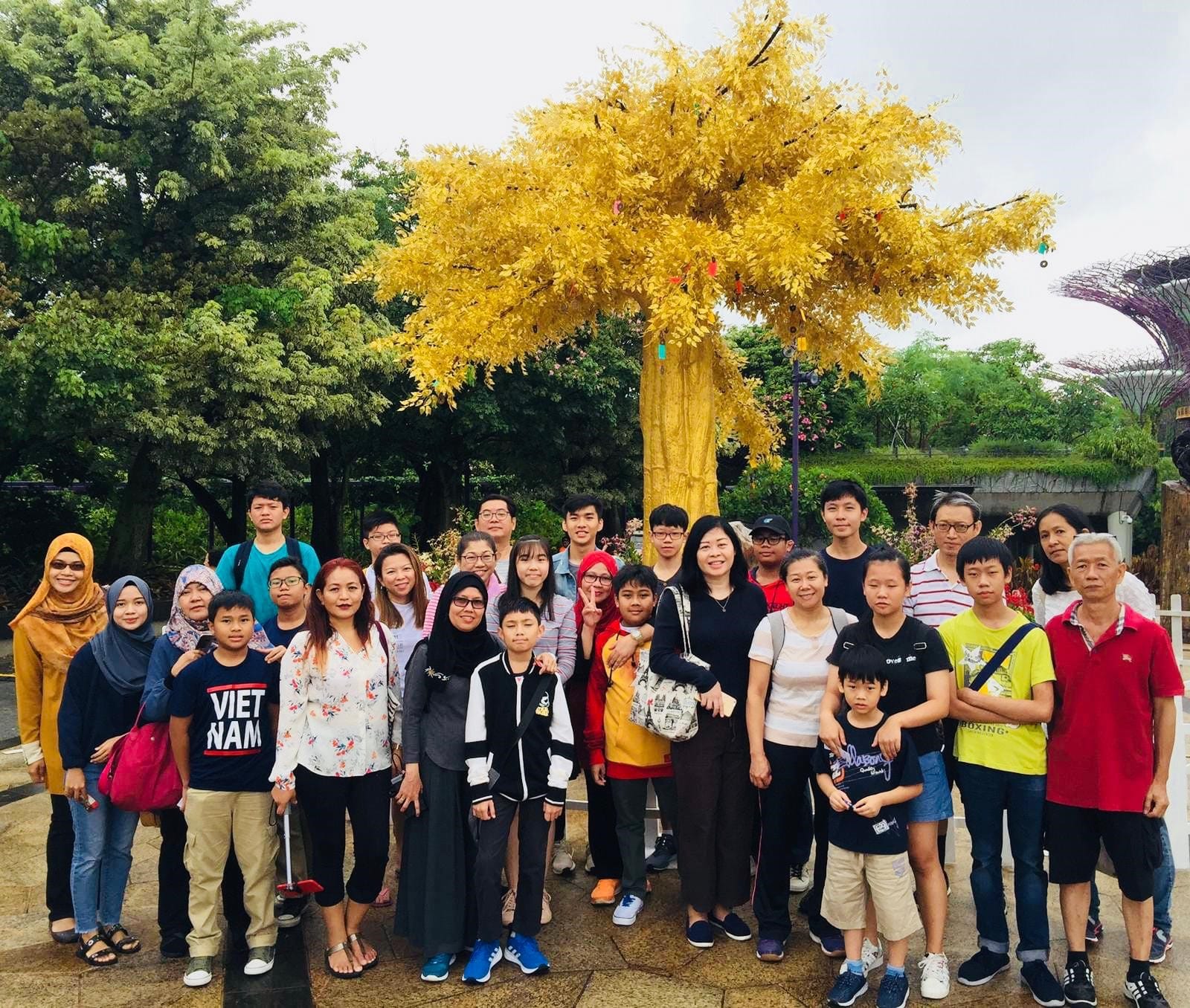 A large group of adults and children pose outdoors in front of a bright yellow-leafed decorative tree in a park.