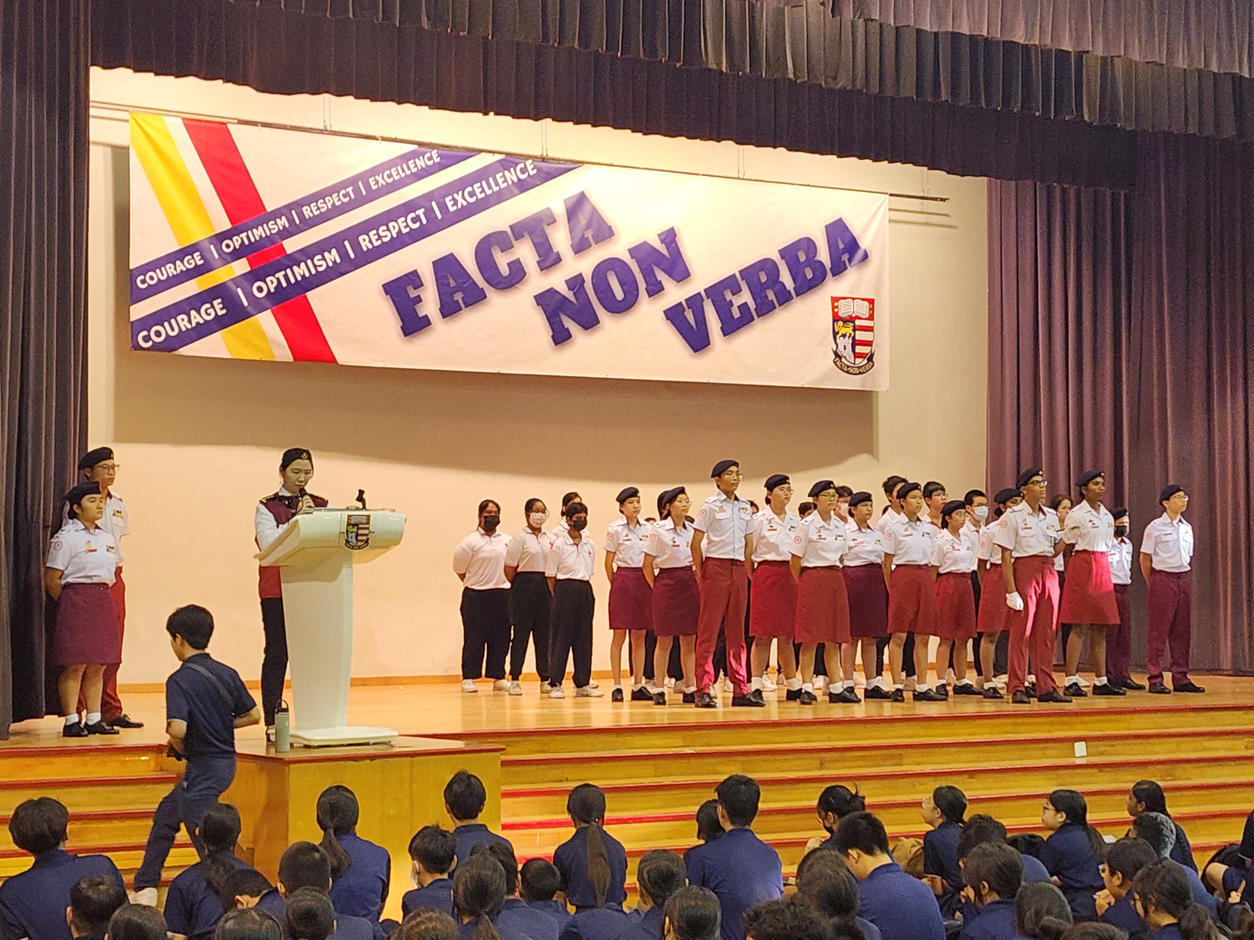 Students in uniform stand on a school stage under a banner reading Facta Non Verba, while an audience watches.