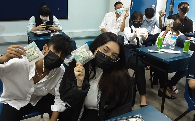 Students wearing black face masks sit at school desks, holding up small green packaged items toward the camera.