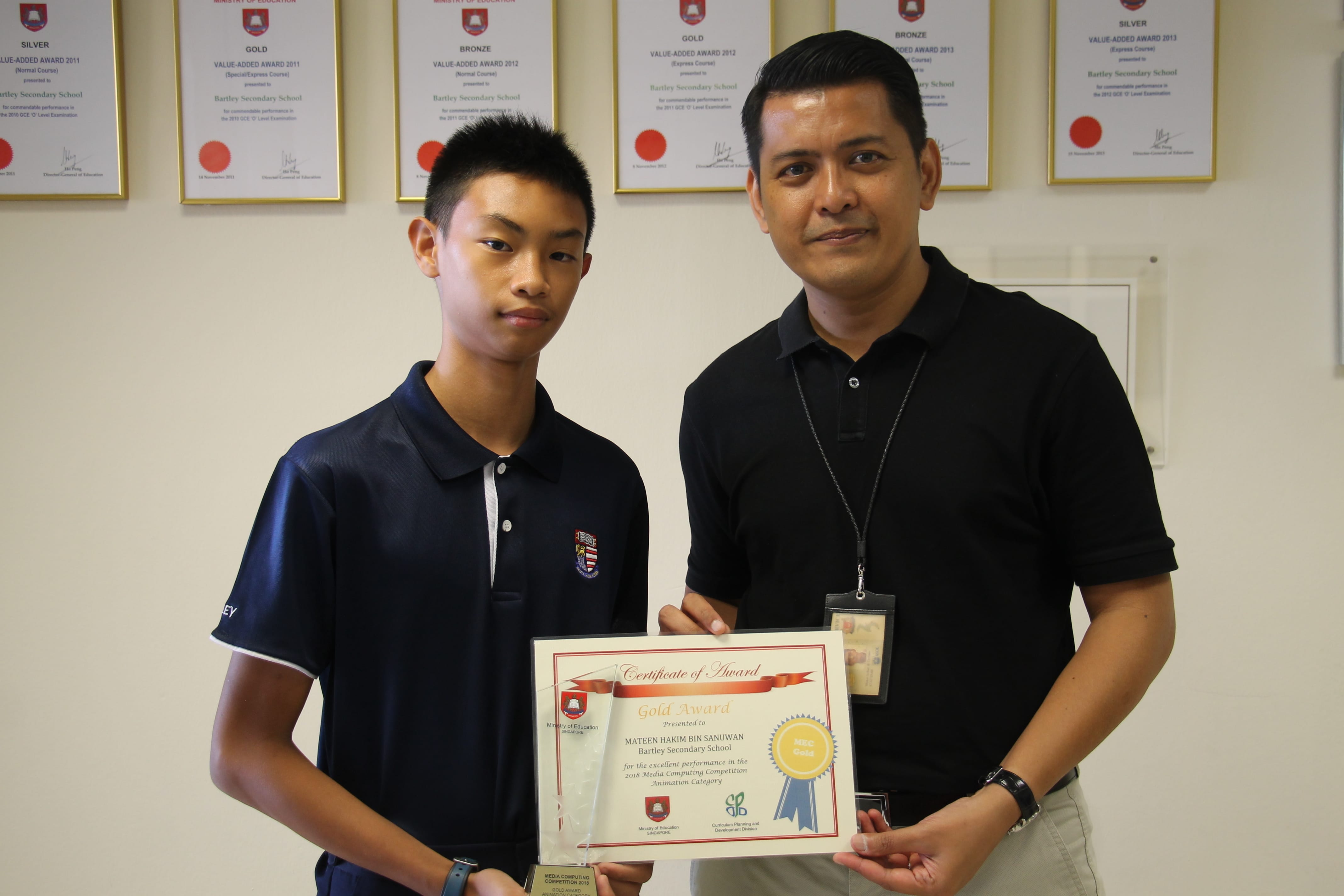 A student and a teacher hold a Gold Award certificate from Bartley Secondary School in front of framed awards.