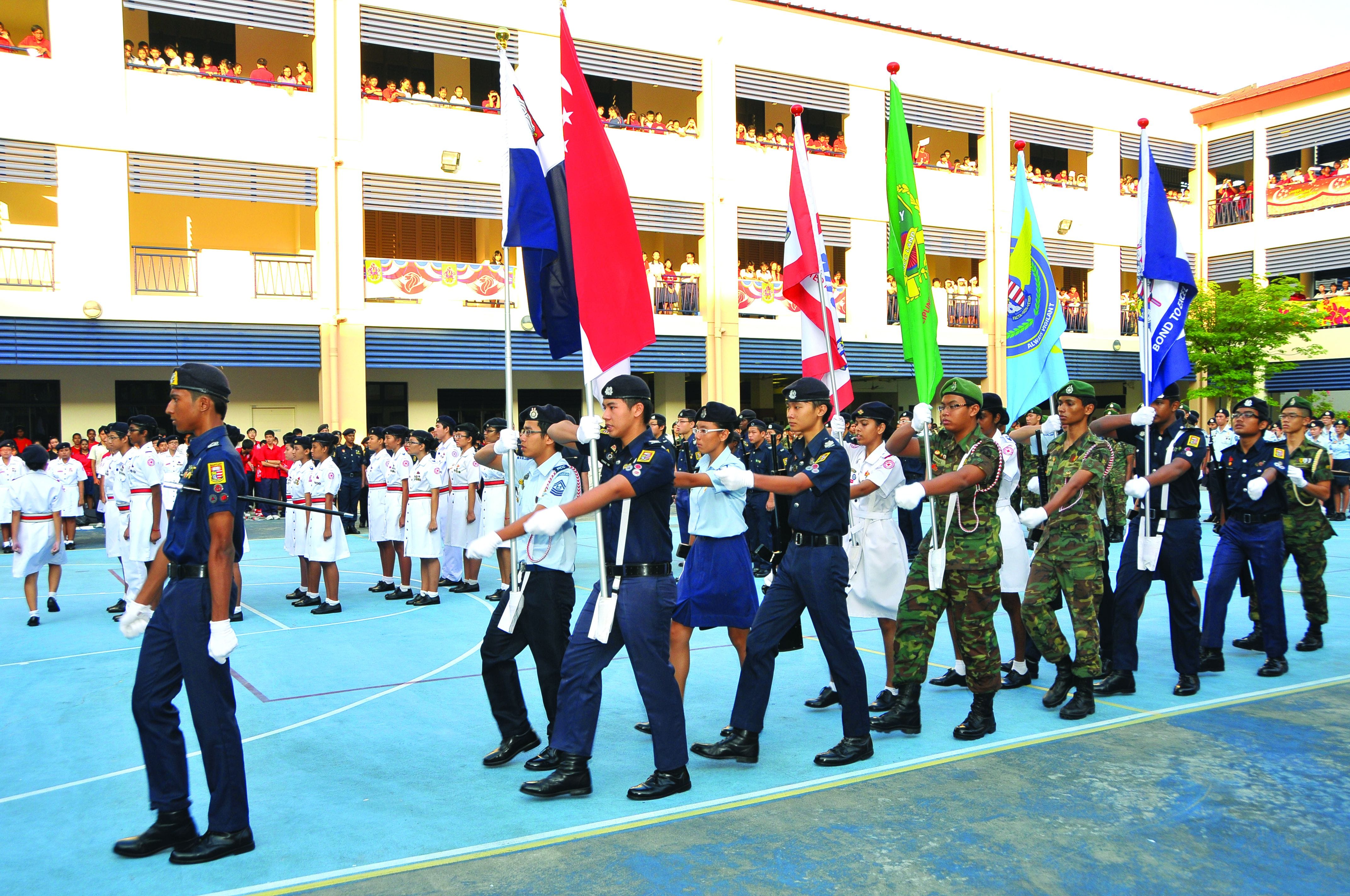 Students in various uniforms march carrying colorful flags in a school courtyard parade, watched by crowds on upper floors.