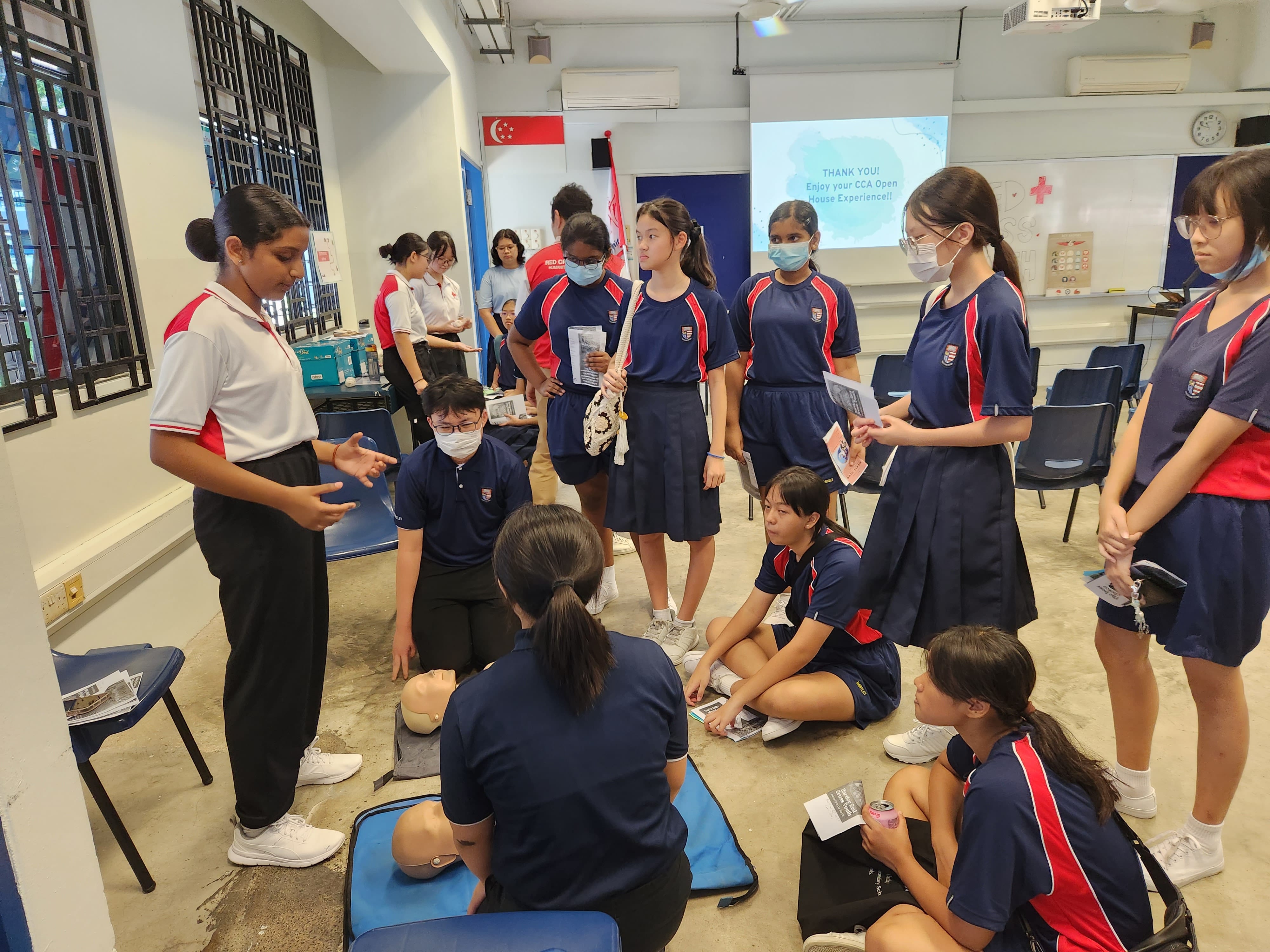 Students in school uniforms gather around a CPR training mannequin during a Red Cross open house demonstration.