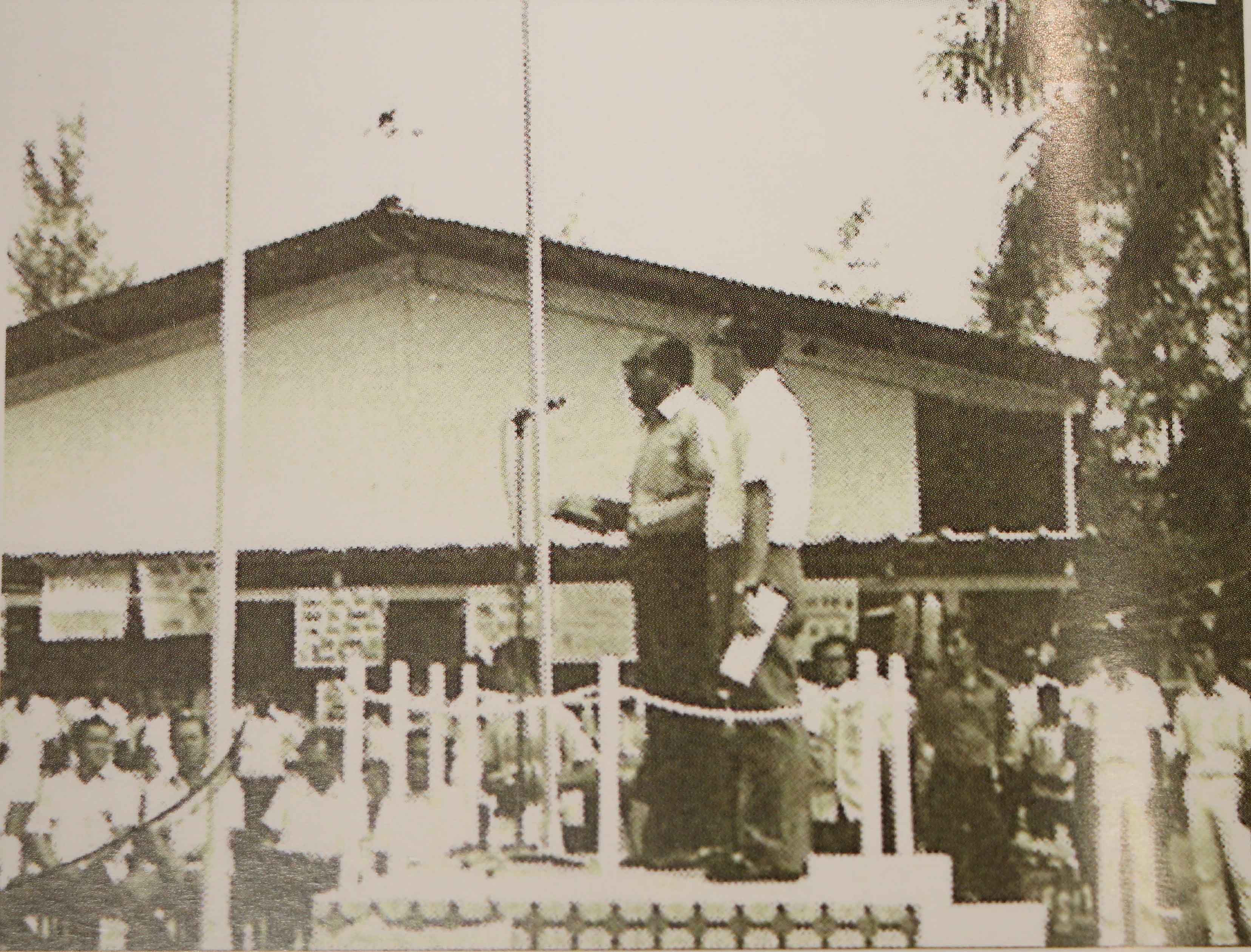 Old sepia photo of two people standing on a raised platform with a microphone, addressed by a crowd outdoors.