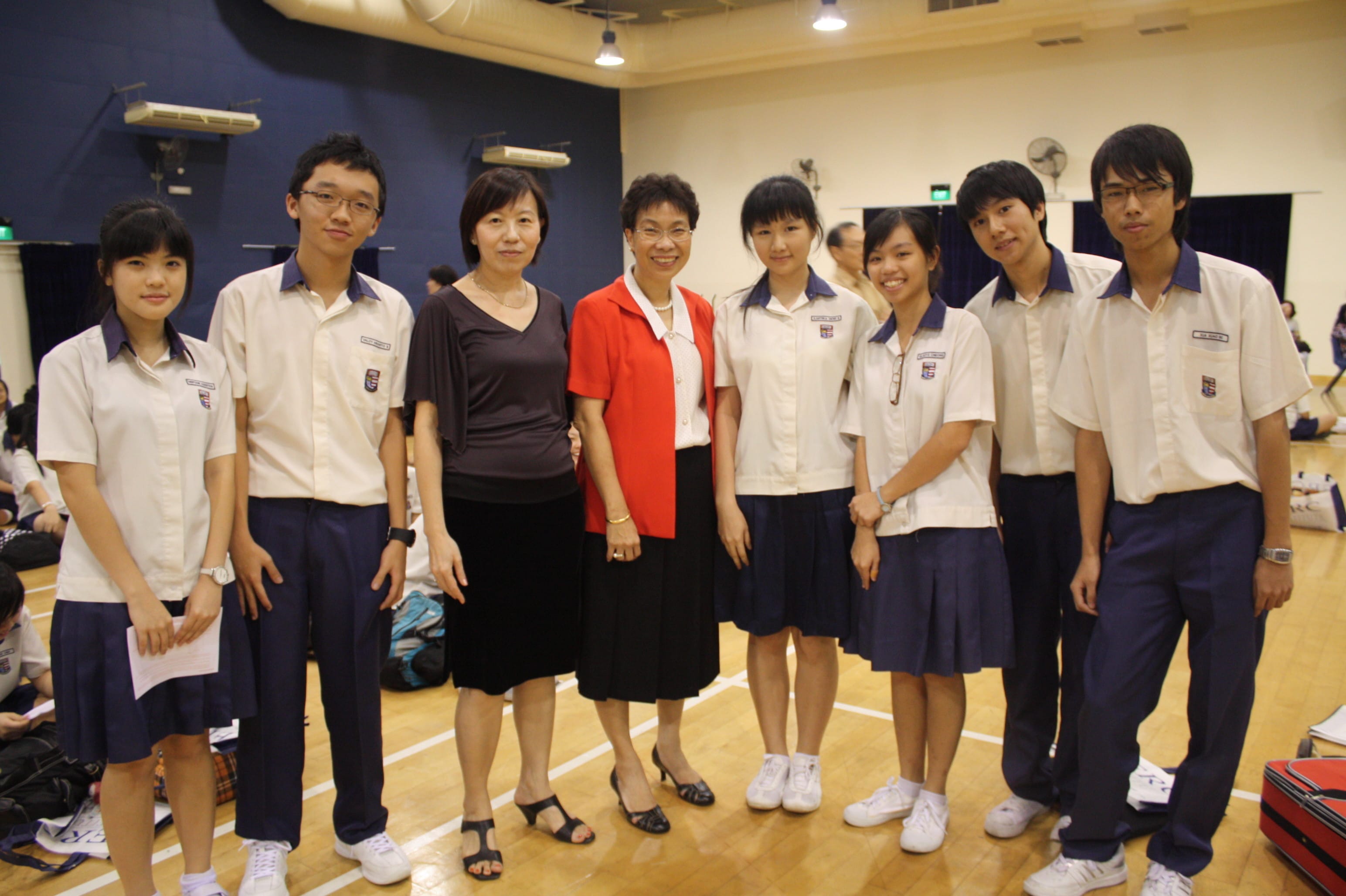Two adults and six students in school uniforms pose together smiling in an indoor gymnasium.