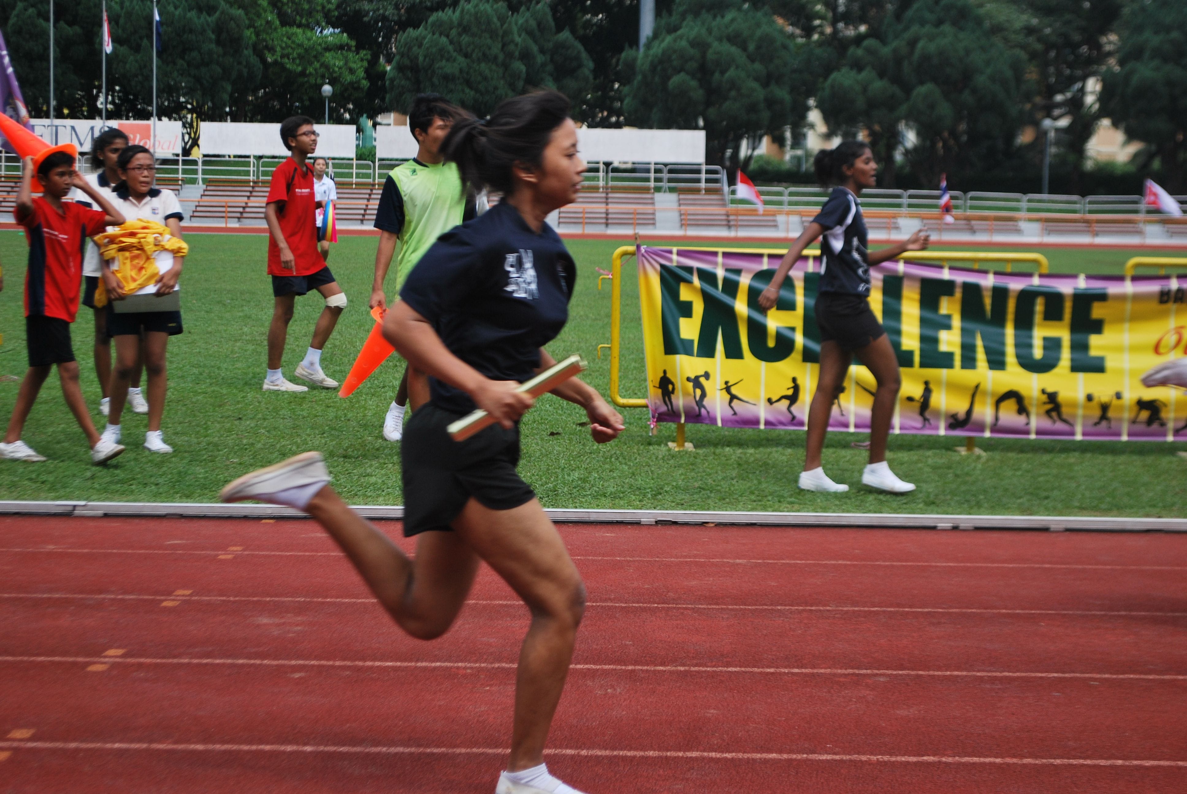 Girl in black sprints on a red track holding a relay baton, with other students and an Excellence banner behind her.