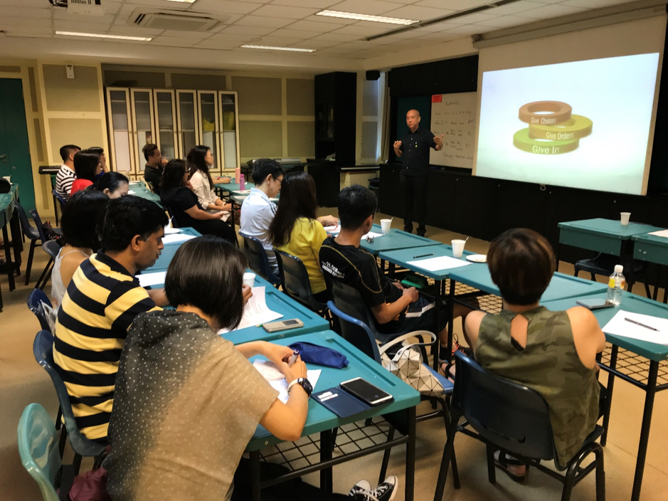A presenter stands before a projected slide showing stacked rings, addressing a classroom of seated adults at teal desks.