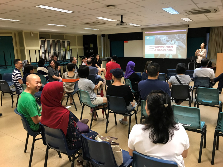 A group of people seated in chairs attend a presentation titled Giving Them a Headstart projected on a screen.