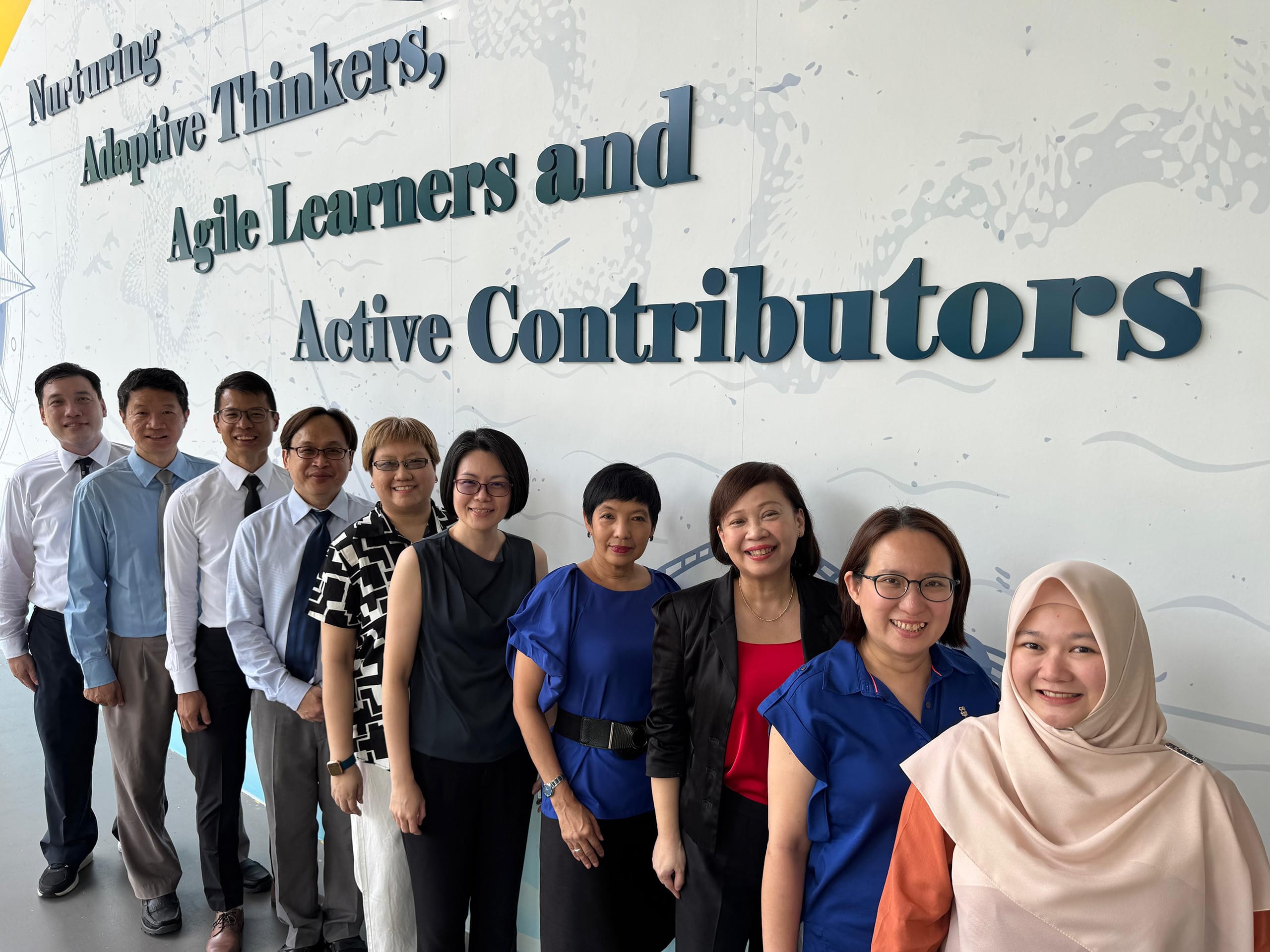 Ten people smiling in a group photo in front of a wall reading Nurturing Adaptive Thinkers, Agile Learners and Active Contributors.