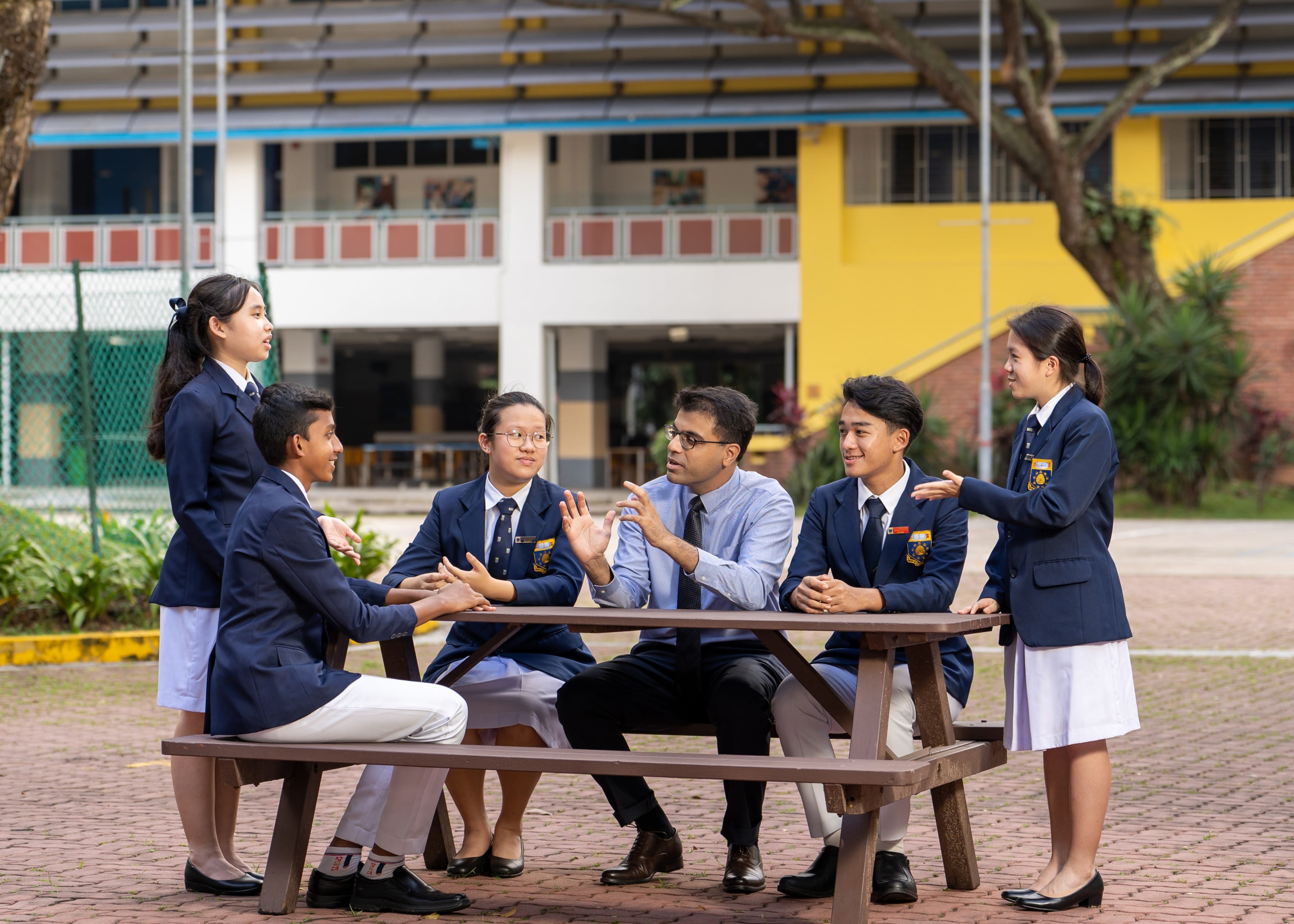 A teacher in a blue shirt discusses with five uniformed students at an outdoor picnic table in a school courtyard.