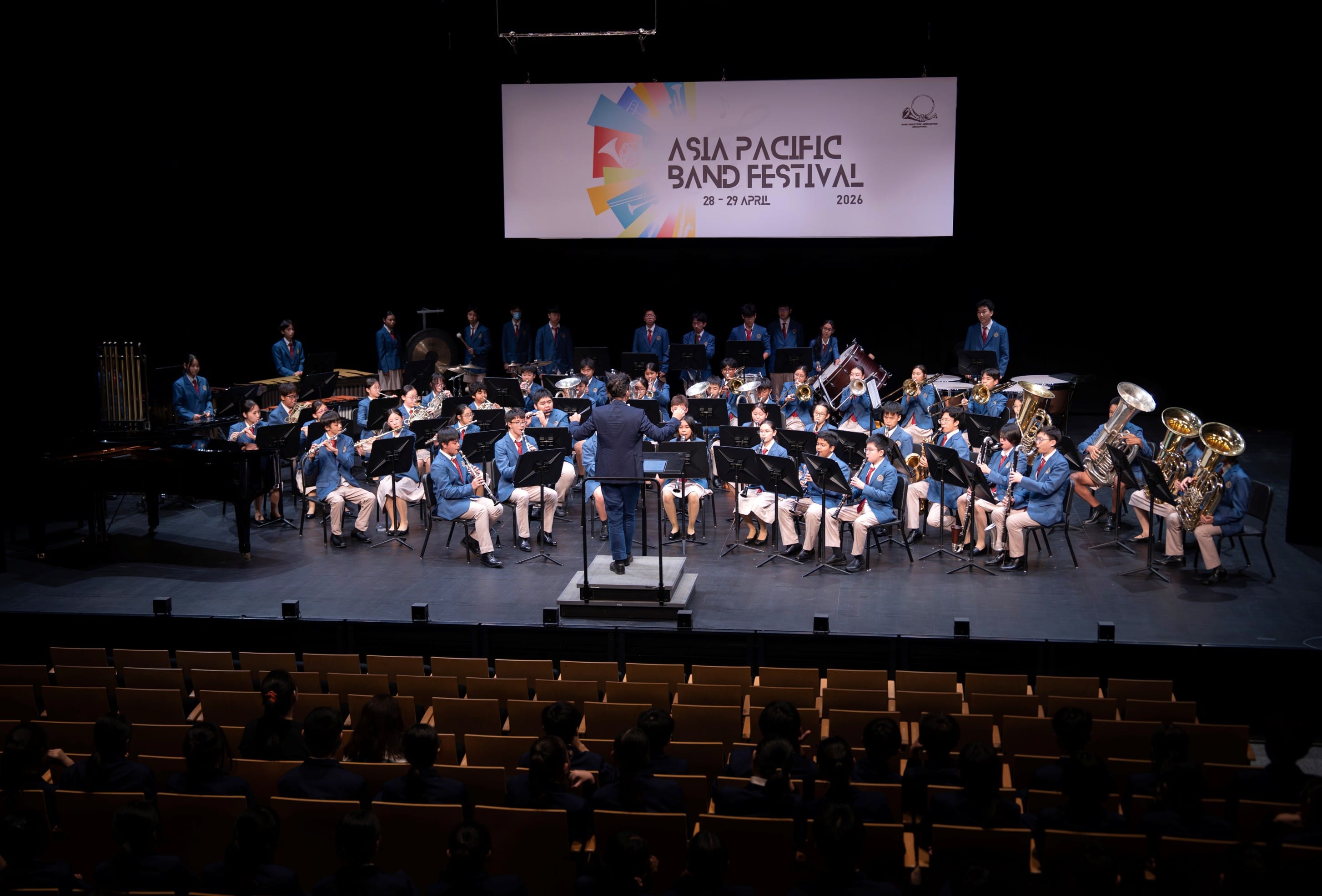 Photograph of the BDS Symphonic Band on stage, performing facing the conductor and a seated audience. A banner “Asia Pacific Band Festival” hangs in the background.