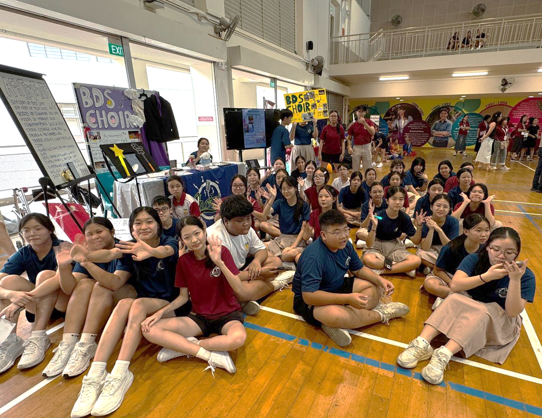 Large group of students seated on the floor in the hall, waving at the camera. They are surrounded by posters promoting BDS Choir.
