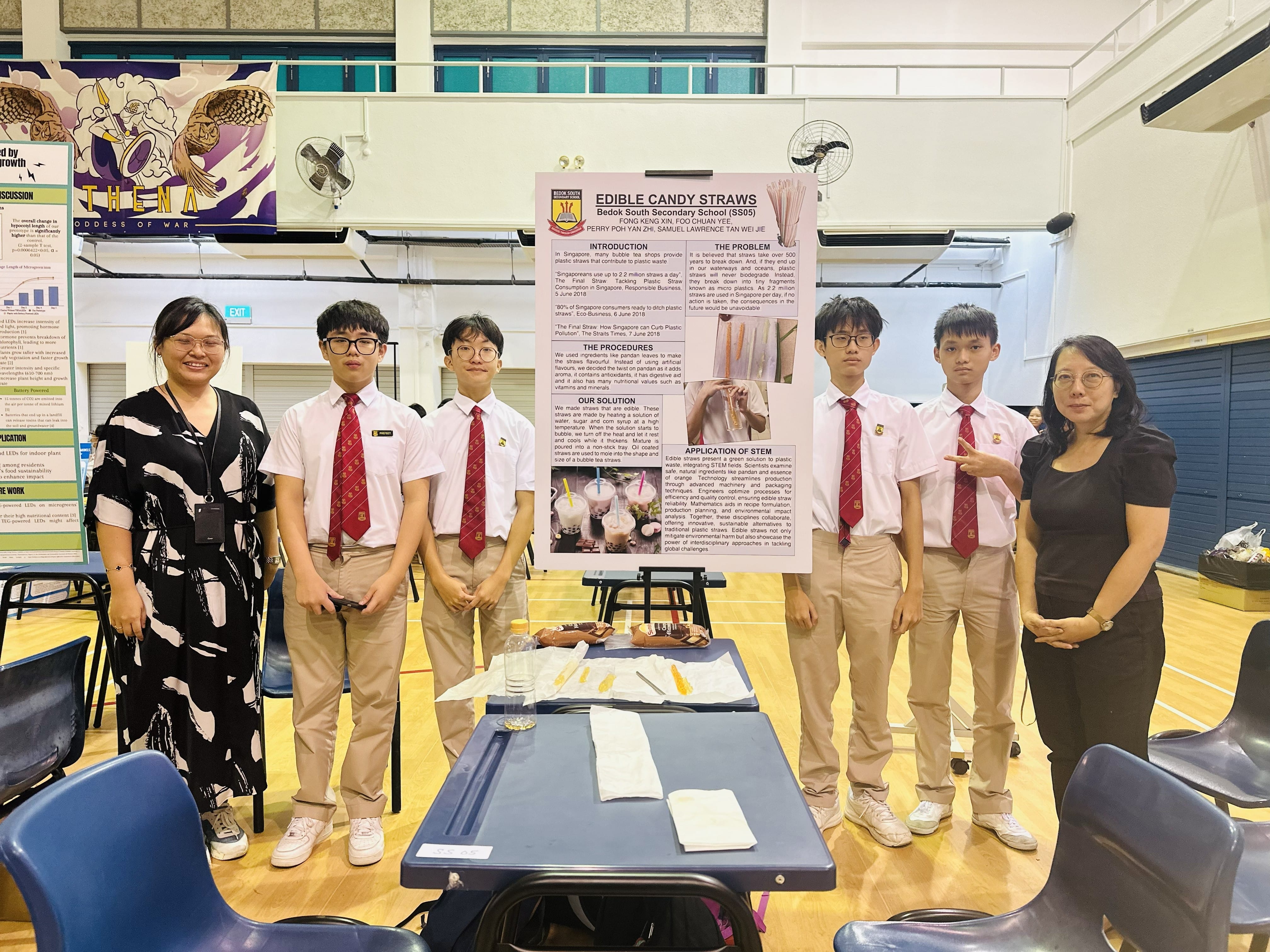 Students and teachers posing next to a large posterboard titled "Candy Straws" in a school hall.