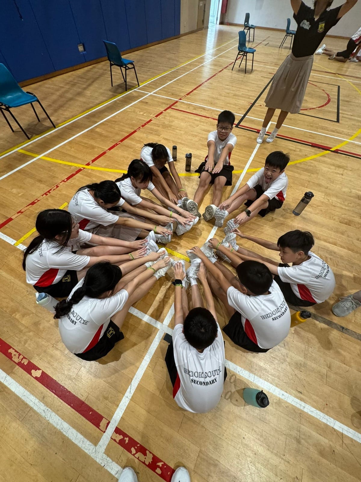 BDS students sit in a circle with extended legs, touching toes together in a synchronized team warm-up exercise.