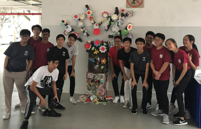 A group of students posing around a colorful tree sculpture made from recycled materials and paper flowers.