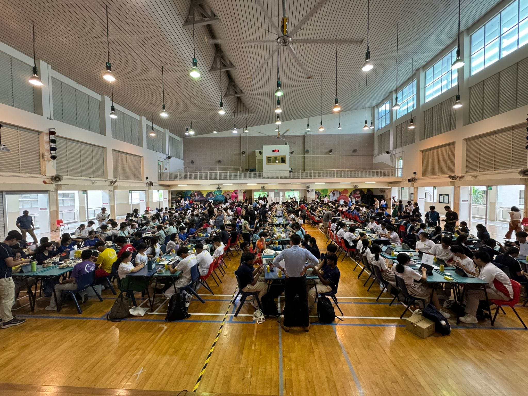 Students wearing different school uniforms sit in long rows of tables inside a large school hall, working on laptops and tablets.