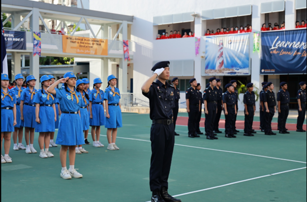Students in blue uniforms and officers in black uniforms stand at salute during a school parade ceremony.