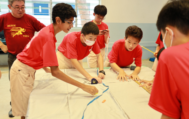 Students in red shirts gather around a table working on a hands-on group activity supervised by an adult.