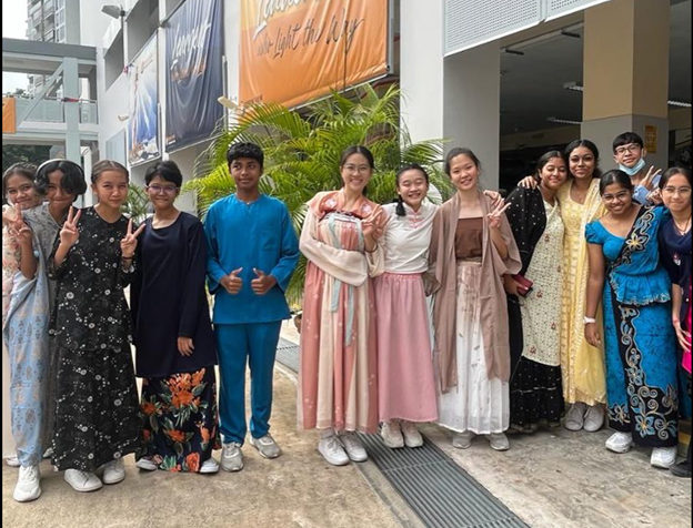 A diverse group of students in traditional cultural attire posing together outdoors near a school building.