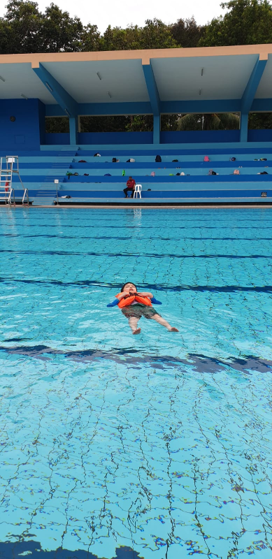 Child wearing an orange life jacket floating on their back in a large outdoor swimming pool with blue bleachers.