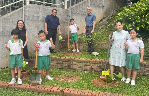 Children in green uniforms and adults pose in a garden area with shovels and a watering can.