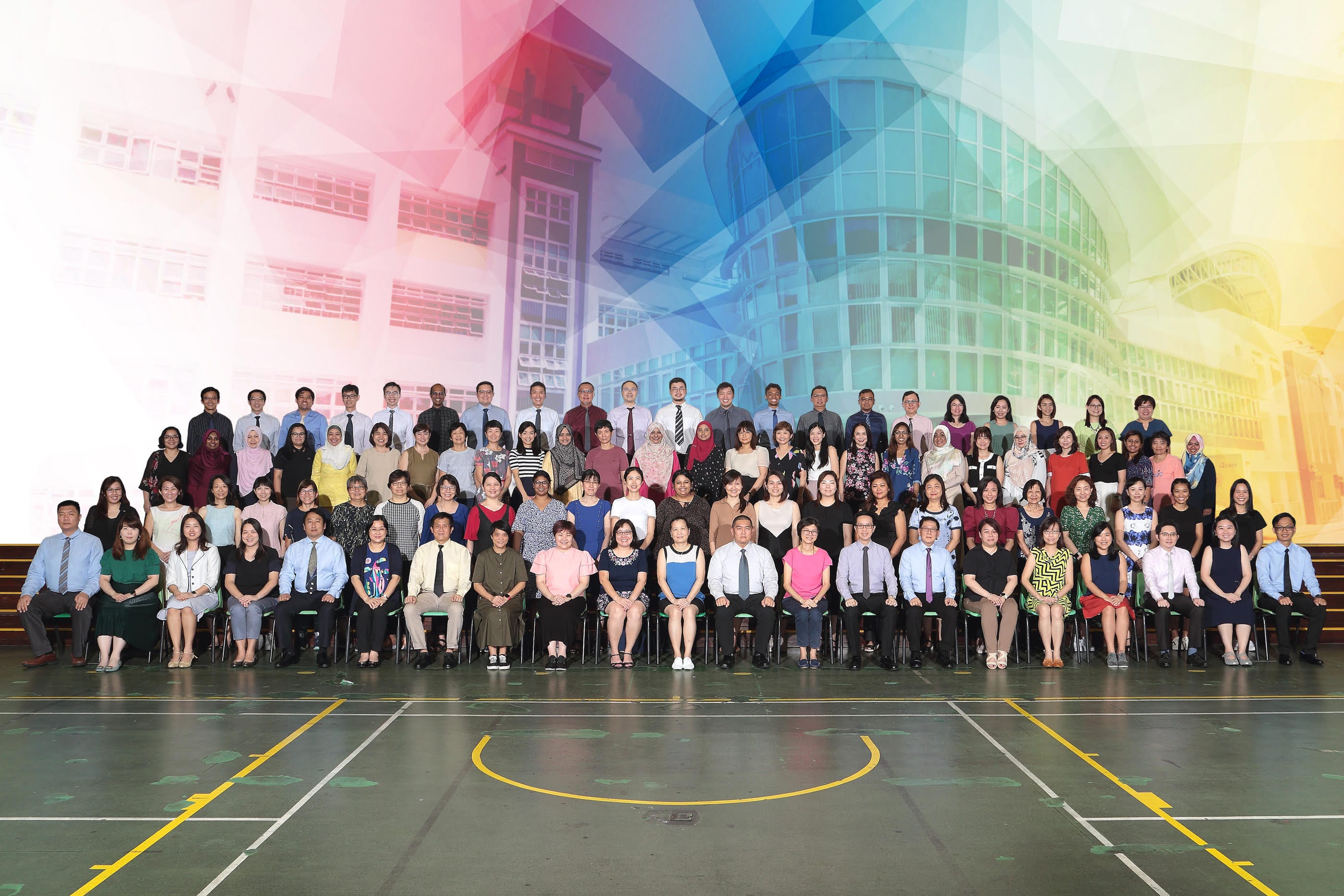 Large group of about 100 people posing in rows on a gymnasium floor before a colorful building backdrop.