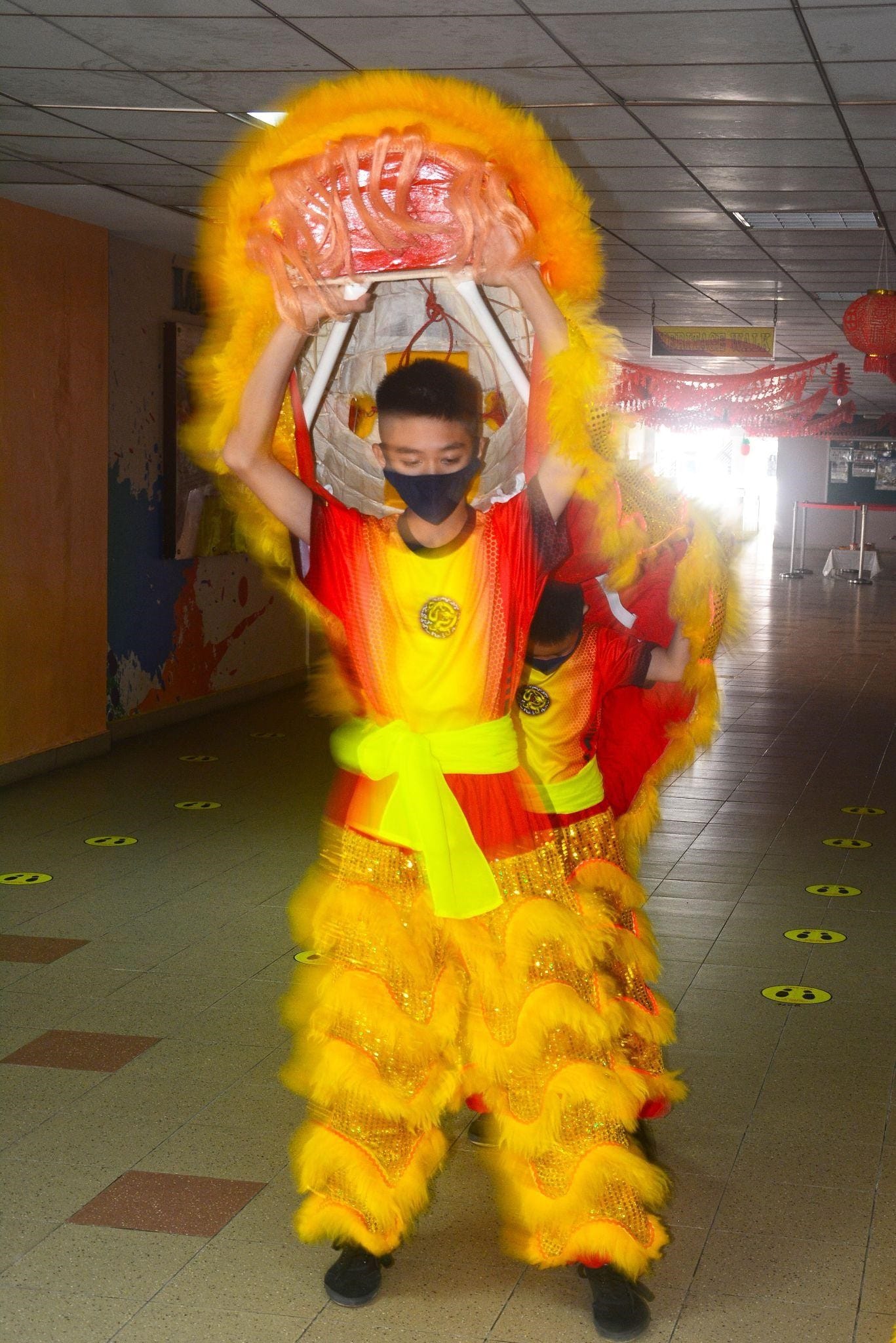 A masked performer in a bright yellow and red costume holds a lion dance head overhead in an indoor hallway.