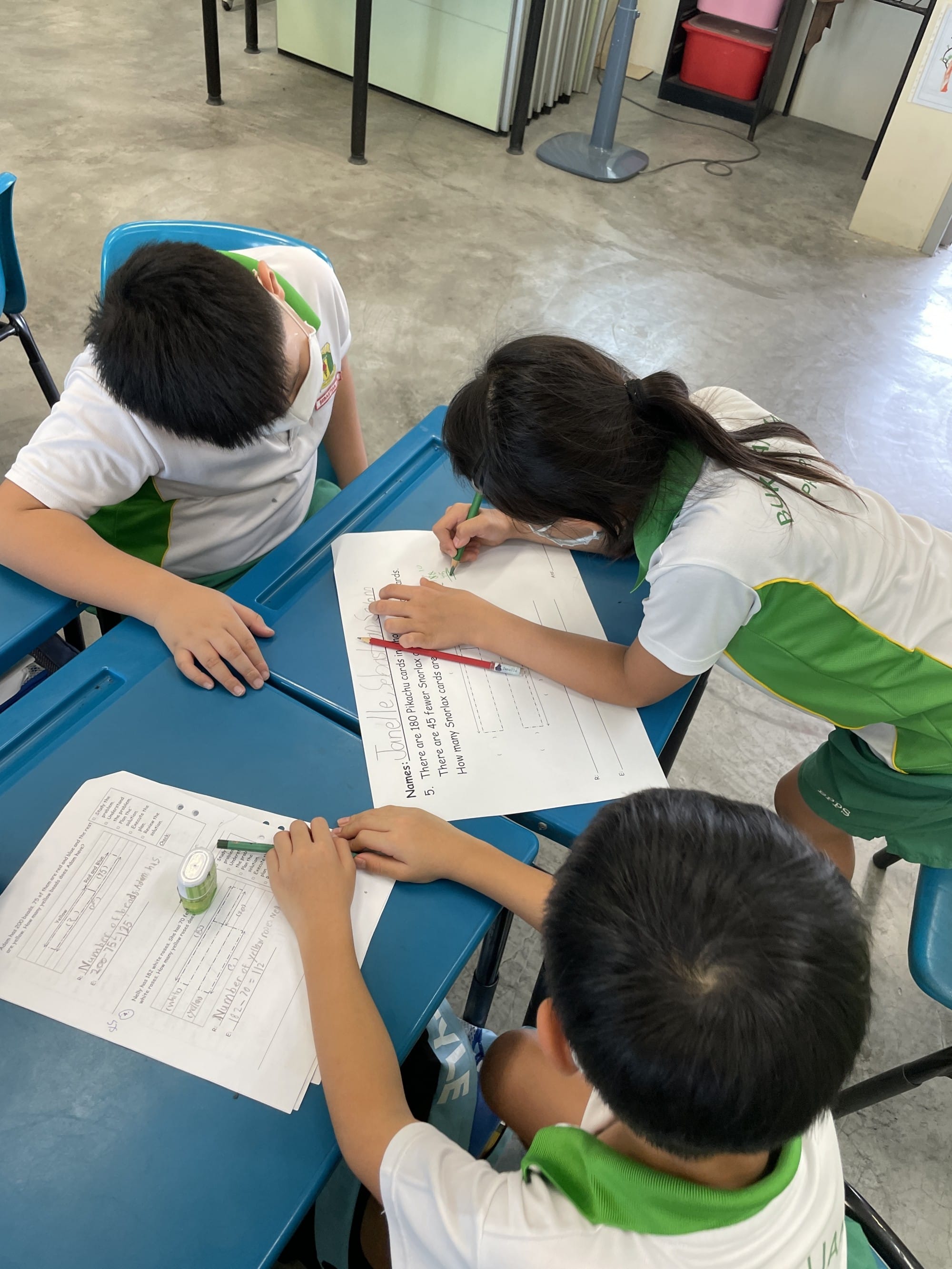Three children in white and green uniforms sit at blue desks, writing on worksheets in a classroom.