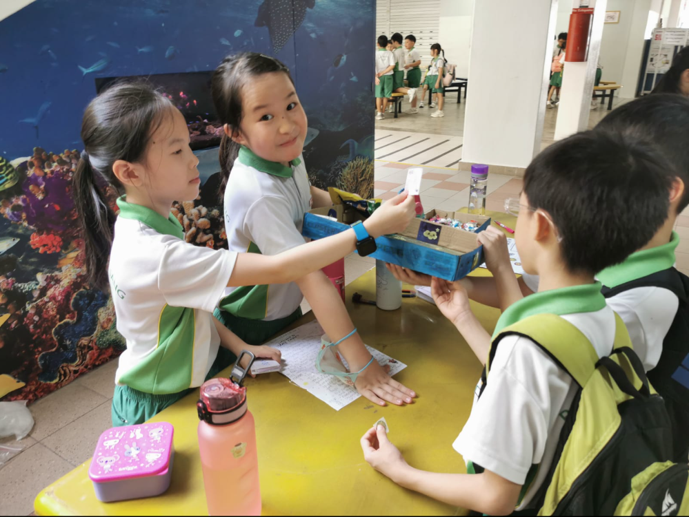 Two schoolgirls in uniforms hand a blue tray of small items to two boys at a table.