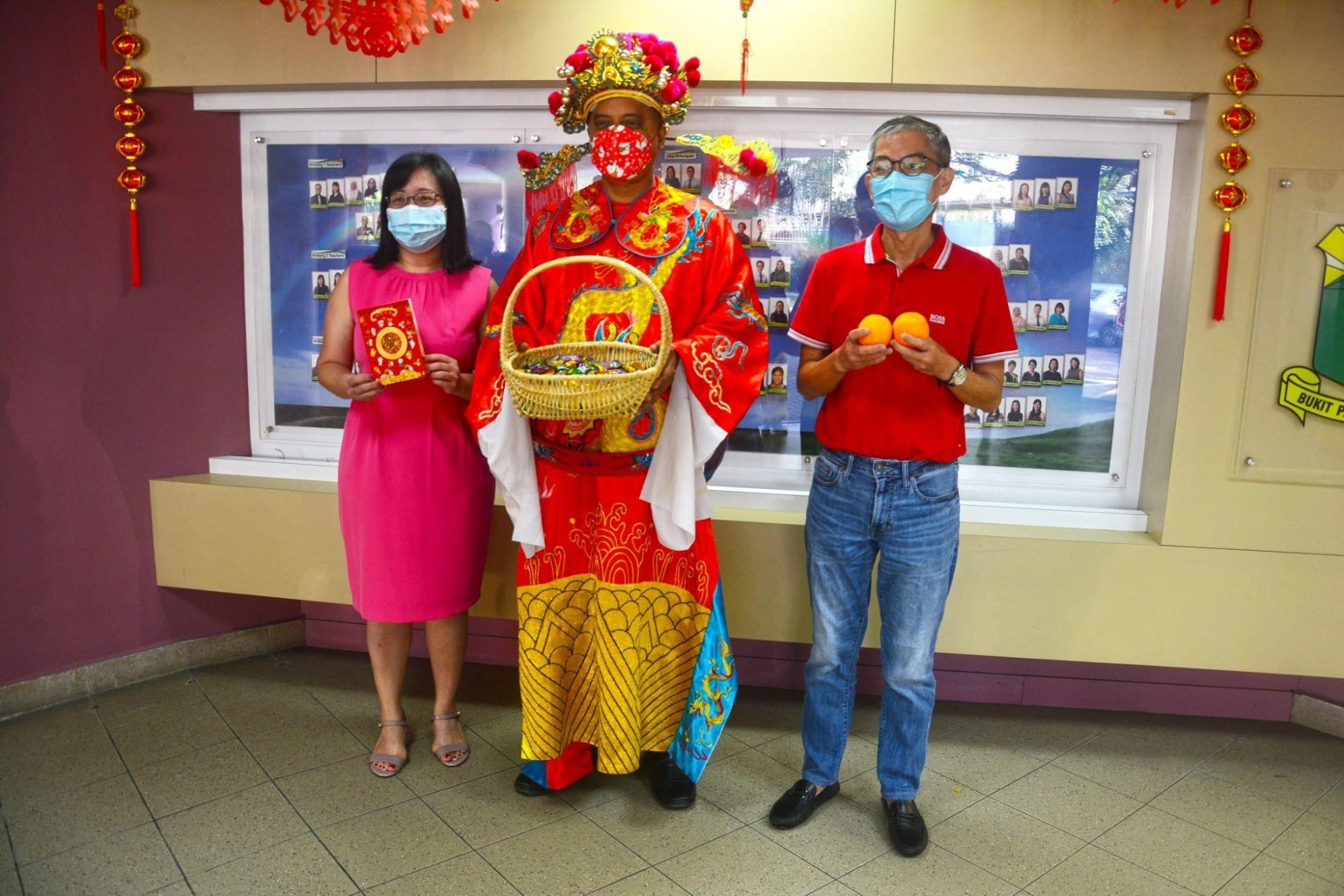 Three masked people pose during Chinese New Year, one in traditional red deity costume holding a basket, flanked by two others.
