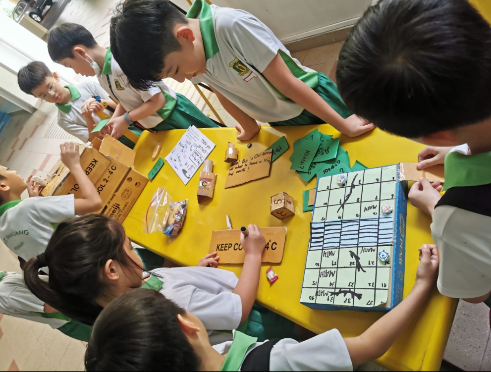 Schoolchildren in green uniforms gather around a yellow table with cardboard signs and a handmade grid board.