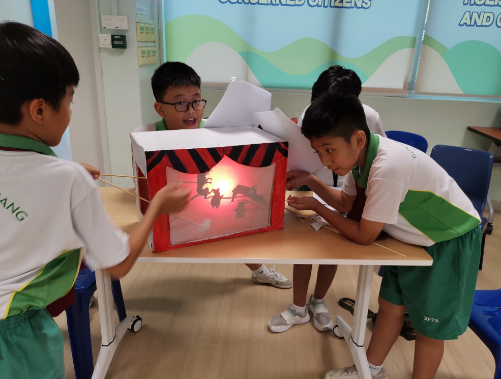 Schoolboys use sticks to make shadow puppets in a lit cardboard theater box on a classroom table.