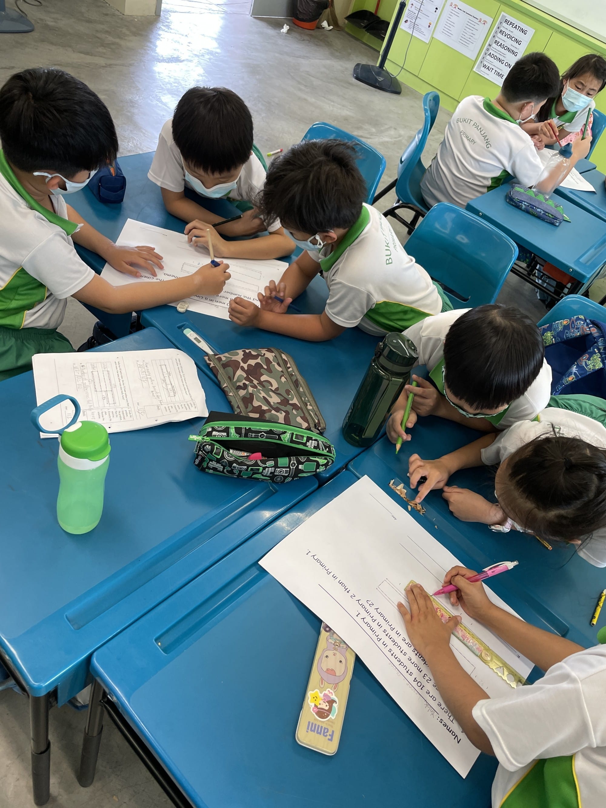 Young students wearing masks work on math worksheets together at blue desks in a primary school classroom.