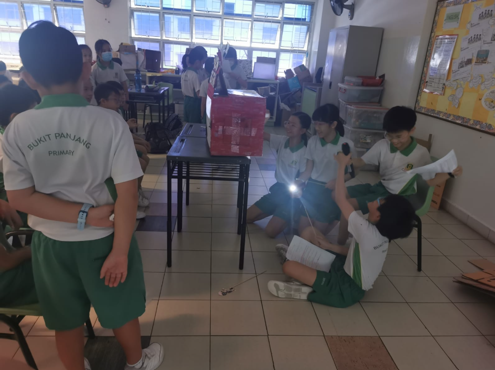 Students in green uniforms gather in a classroom around tables; one holds a flashlight toward a red box.