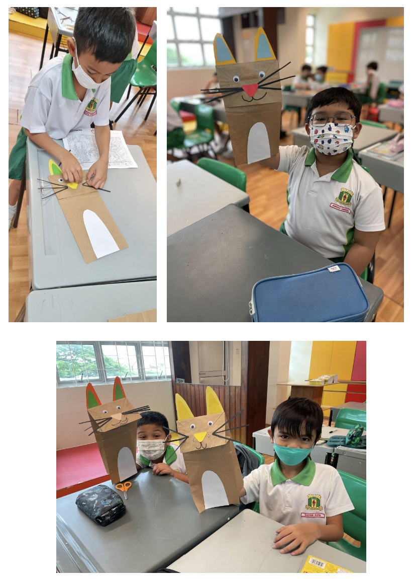 Three masked school children display handmade cardboard cat puppets with googly eyes and whiskers at classroom desks.