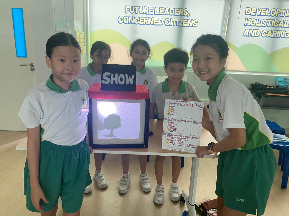 Five students in white-and-green uniforms pose by a SHOW shadow puppet box; one holds a quiz board.