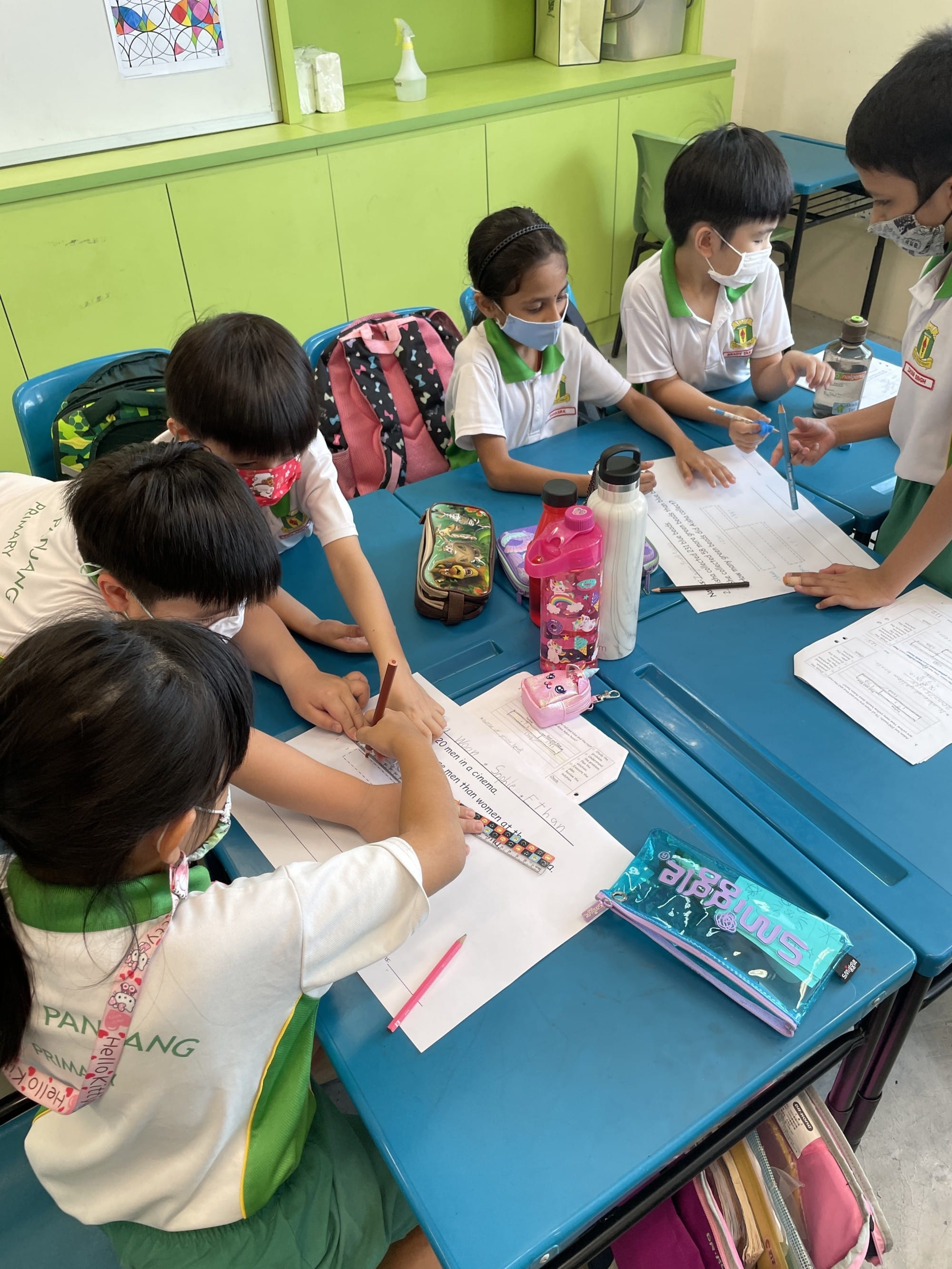 Five young students in school uniforms and masks sit around a blue table, writing on worksheets in a classroom.