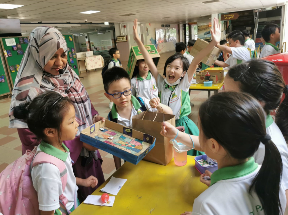 Schoolchildren around a table as an adult in a headscarf watches; one girl cheers with both arms raised.