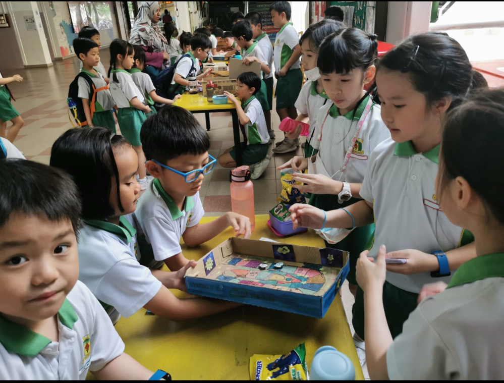 Schoolchildren in green uniforms gather around tables, showing a cardboard game board and snacks.