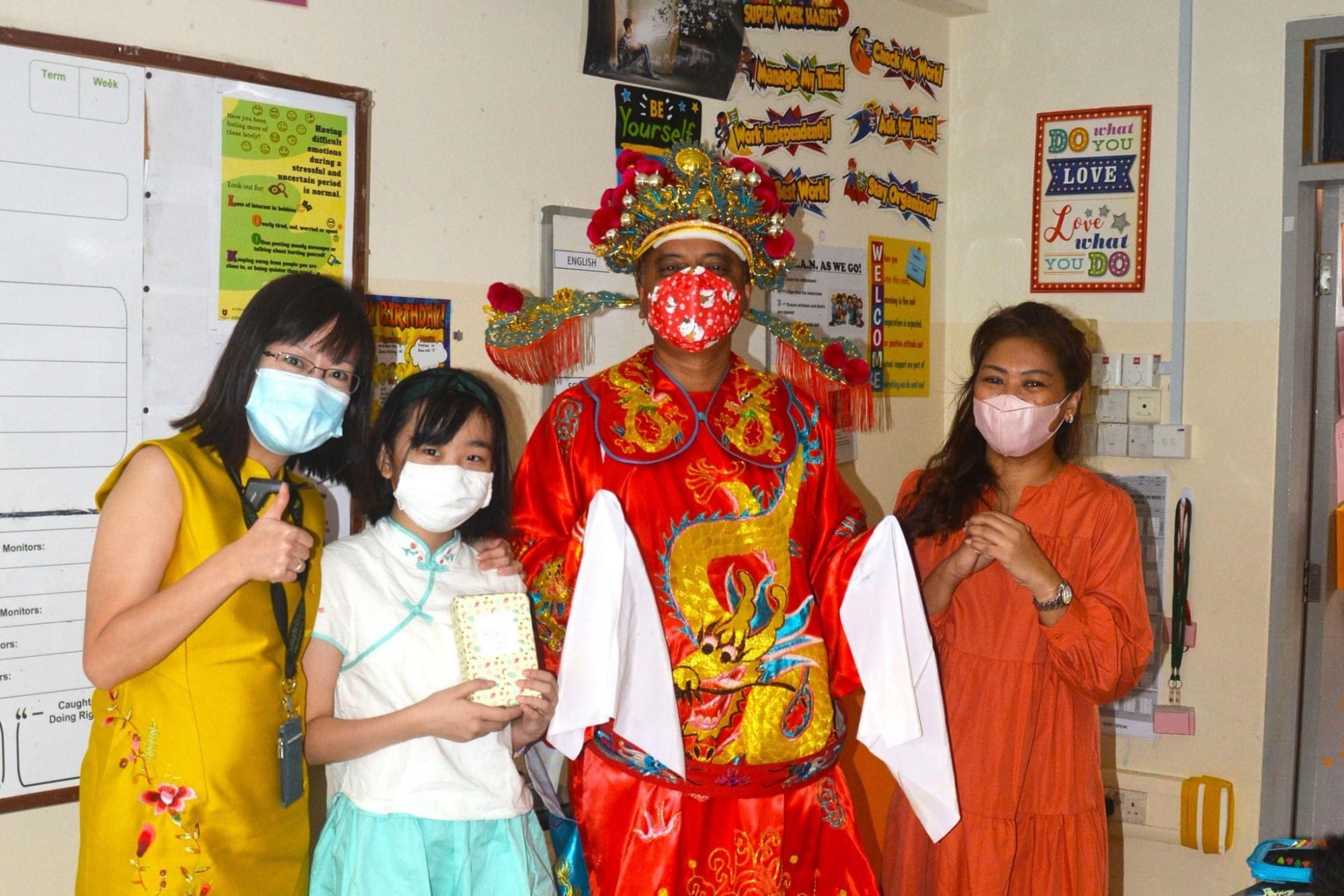 Four masked people pose in a classroom, one wearing a red Chinese emperor costume with ornate gold crown.
