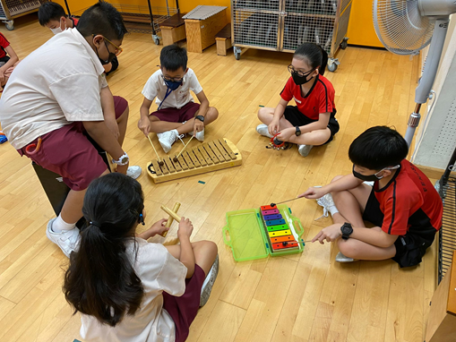 Five masked children sit on a wooden floor playing xylophones, with an adult supervising nearby.