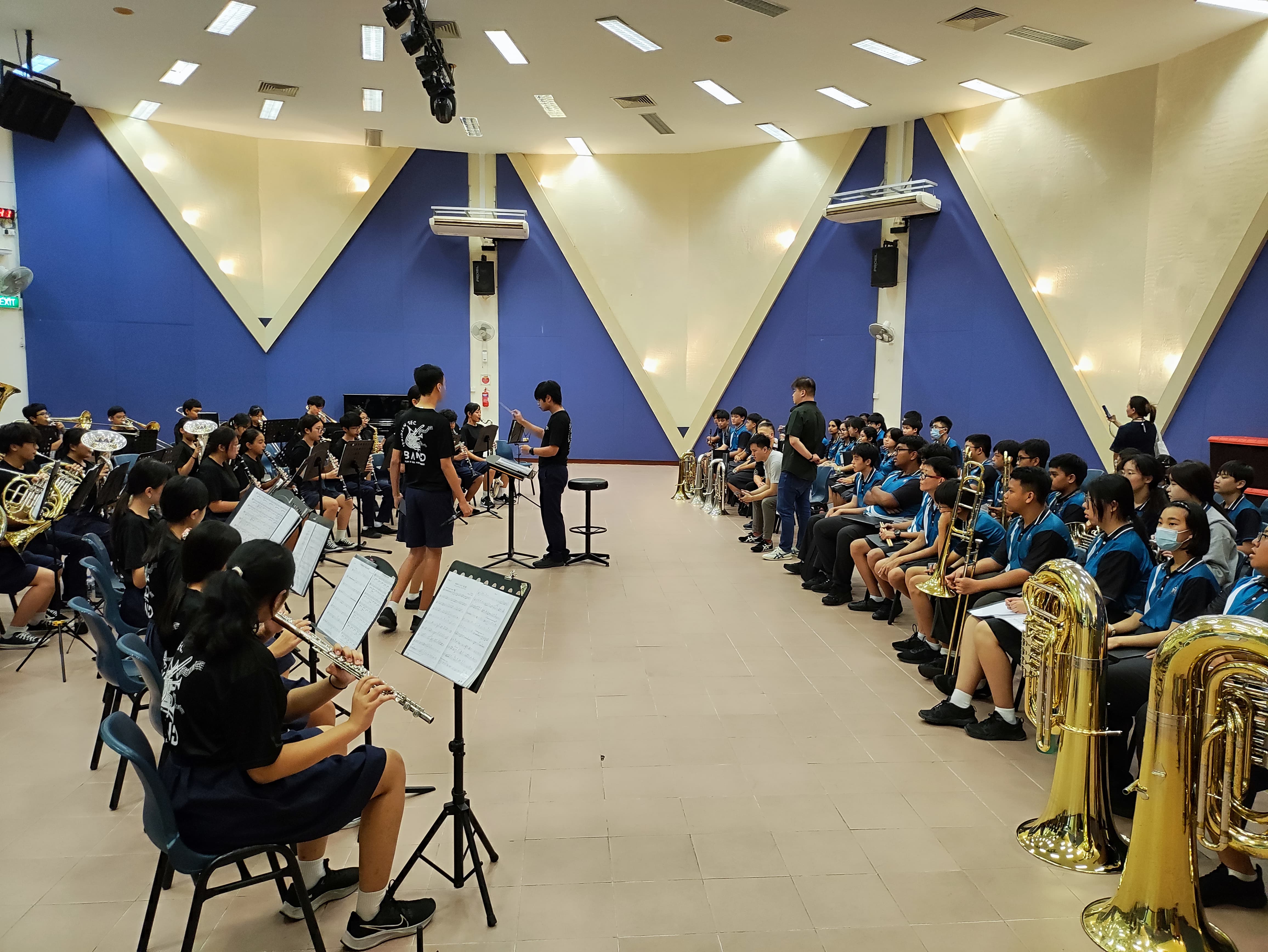Students in a band rehearsal room play brass and wind instruments while a conductor stands at a music stand.
