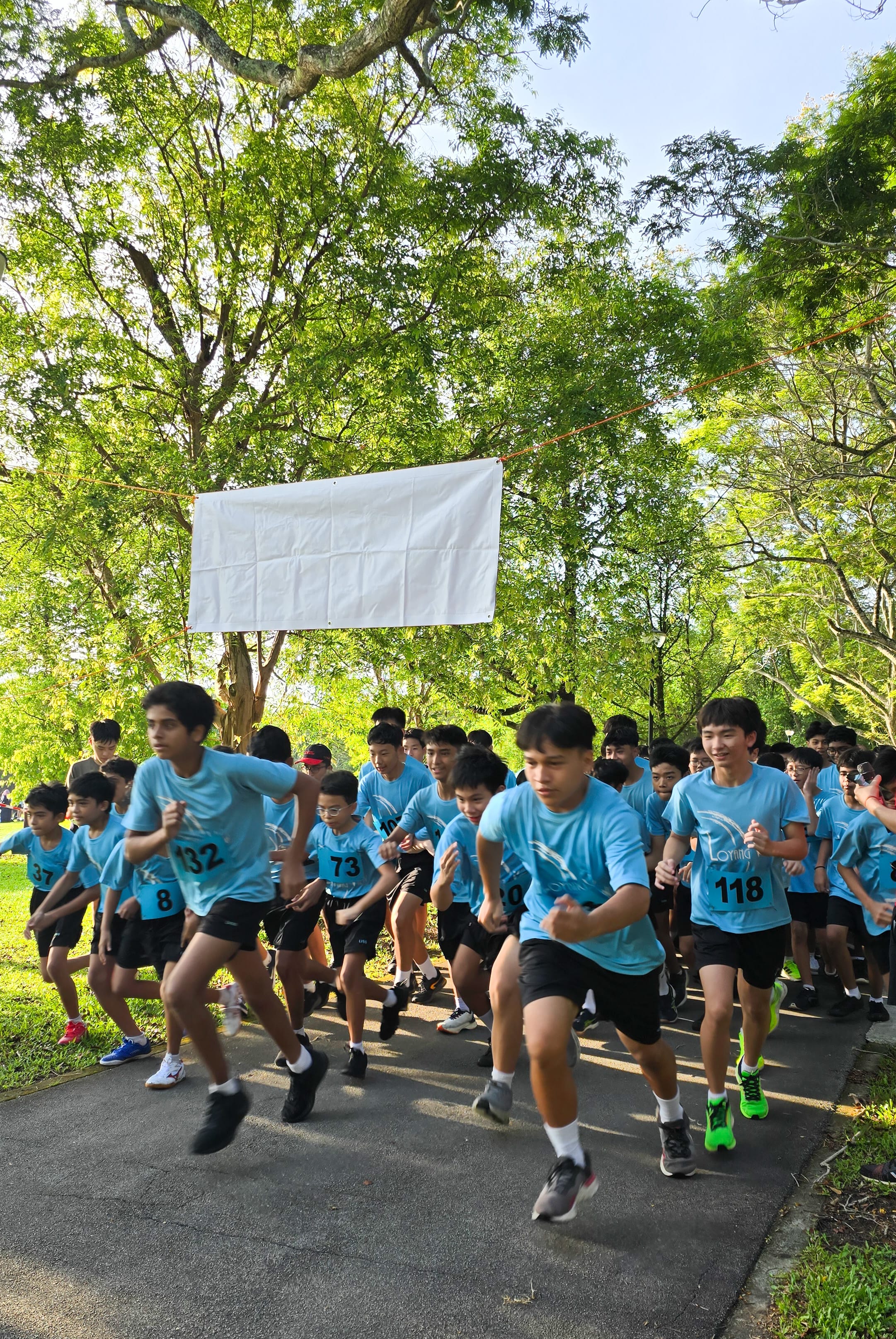 B division students energetically sprint forward from a starting line during an outdoor race. A blank white banner hangs suspended between trees above the path, and a lush green forest fills the background.