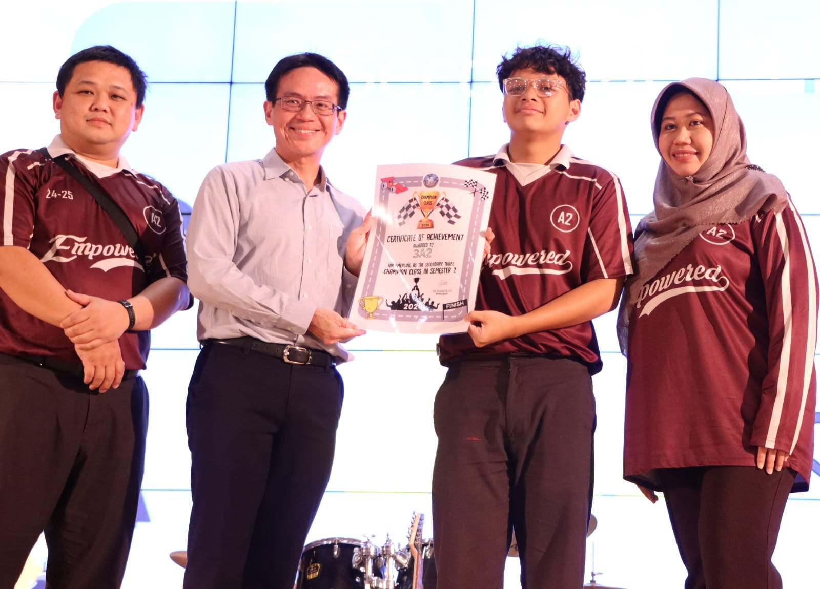 Four people stand together as a man in a blue shirt presents a Certificate of Achievement to a student.