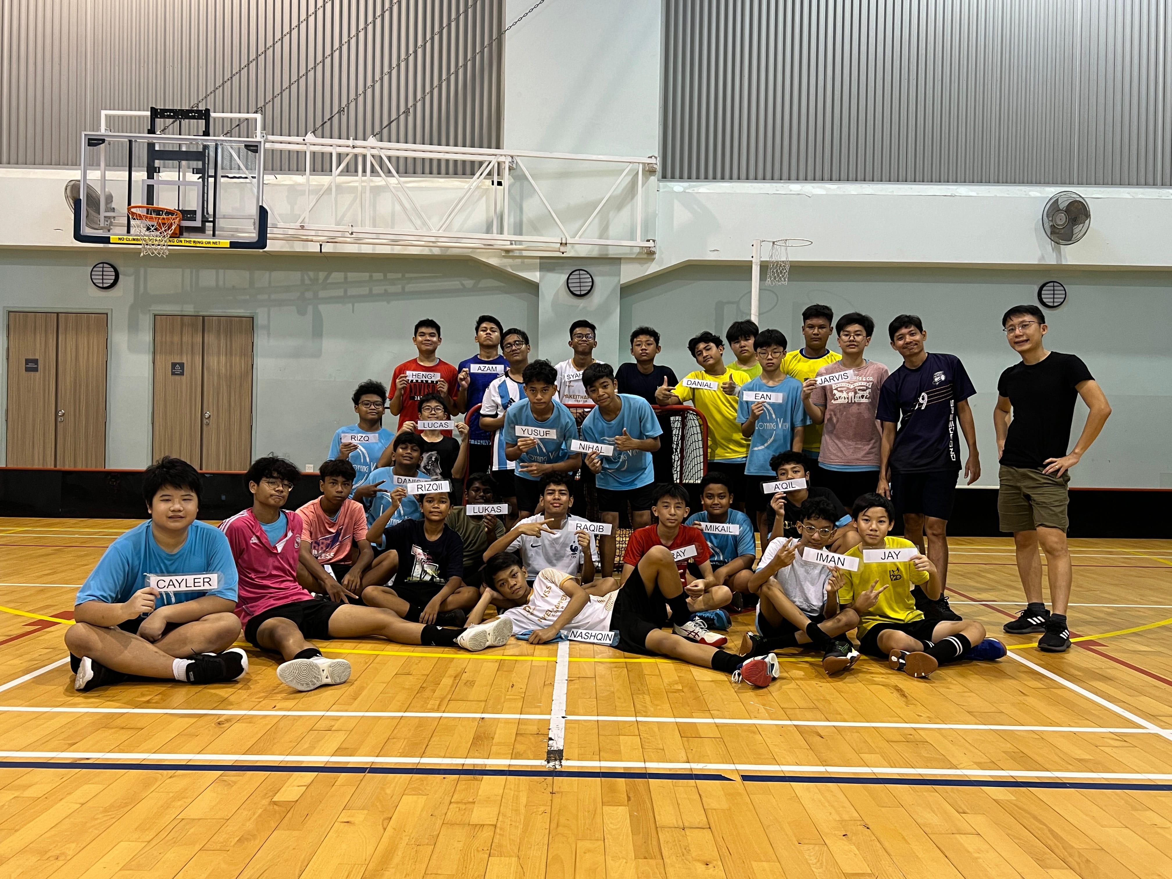 A large group of young boys holding name cards pose together on an indoor basketball court.