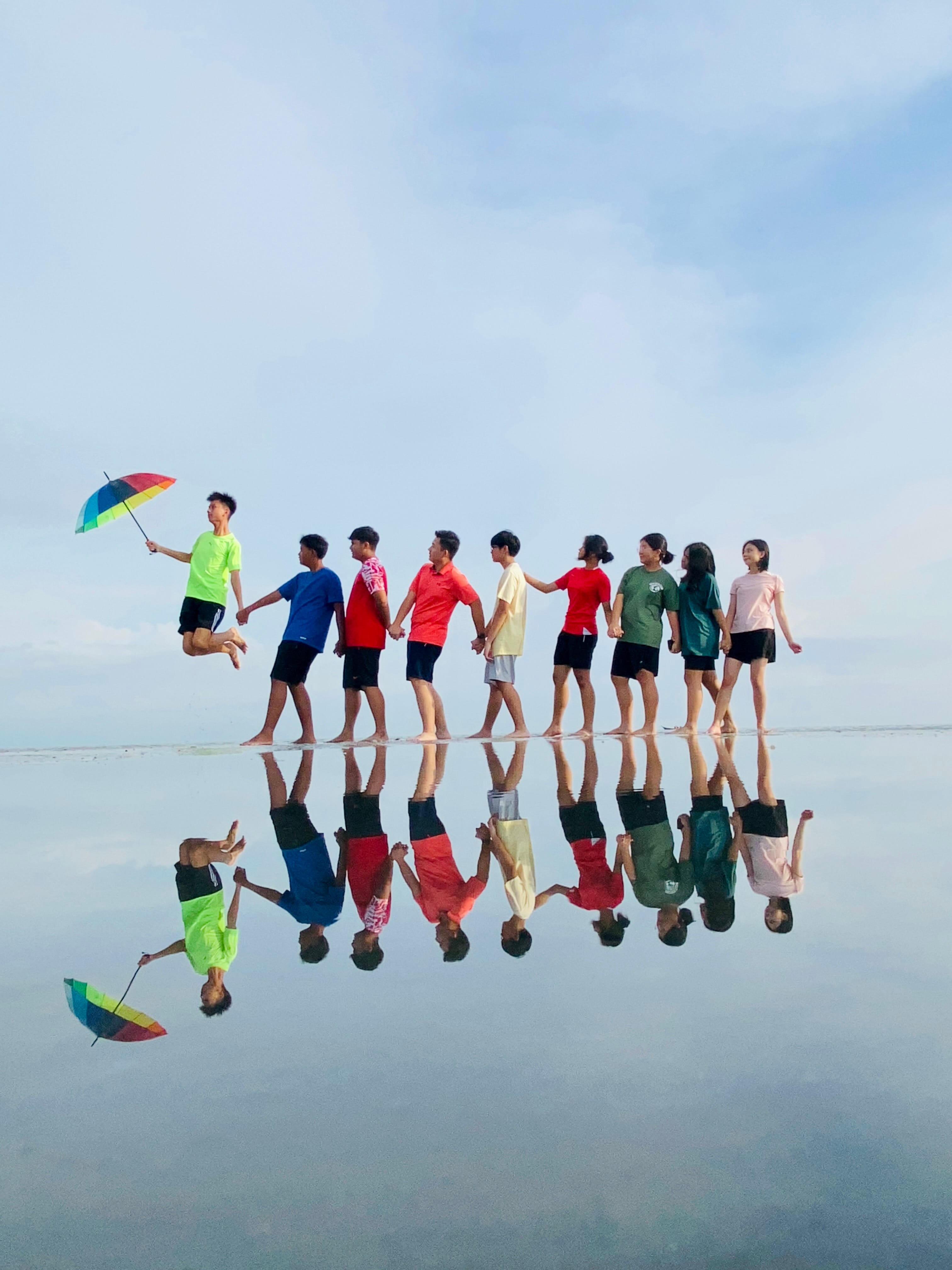Nine people in colorful shirts hold hands in a line on a reflective wet beach, one jumping with a rainbow umbrella.