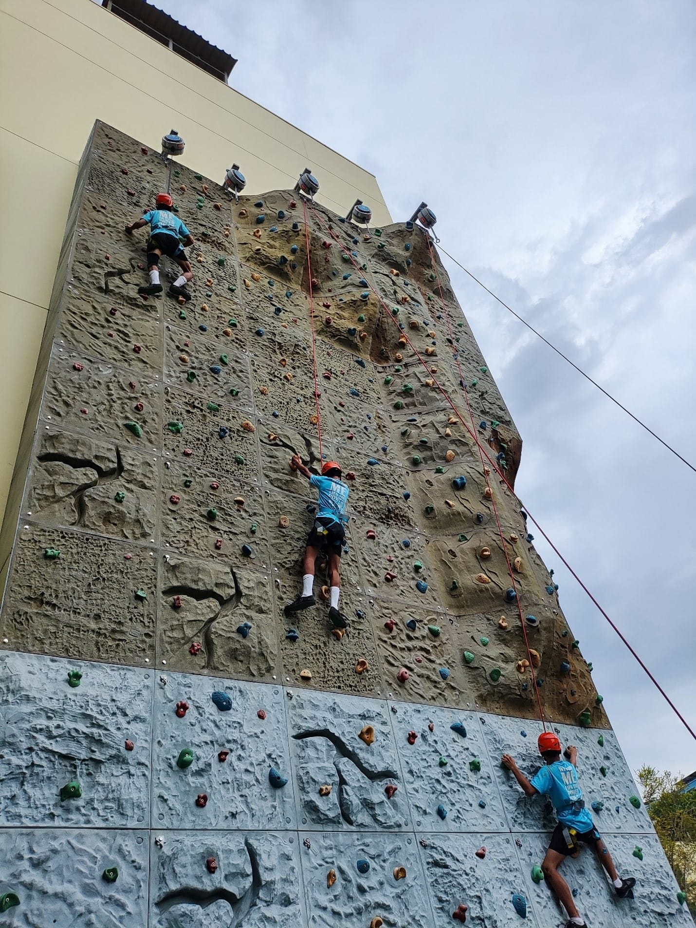 Three children in blue shirts and red helmets climb a tall outdoor rock climbing wall with colorful holds.